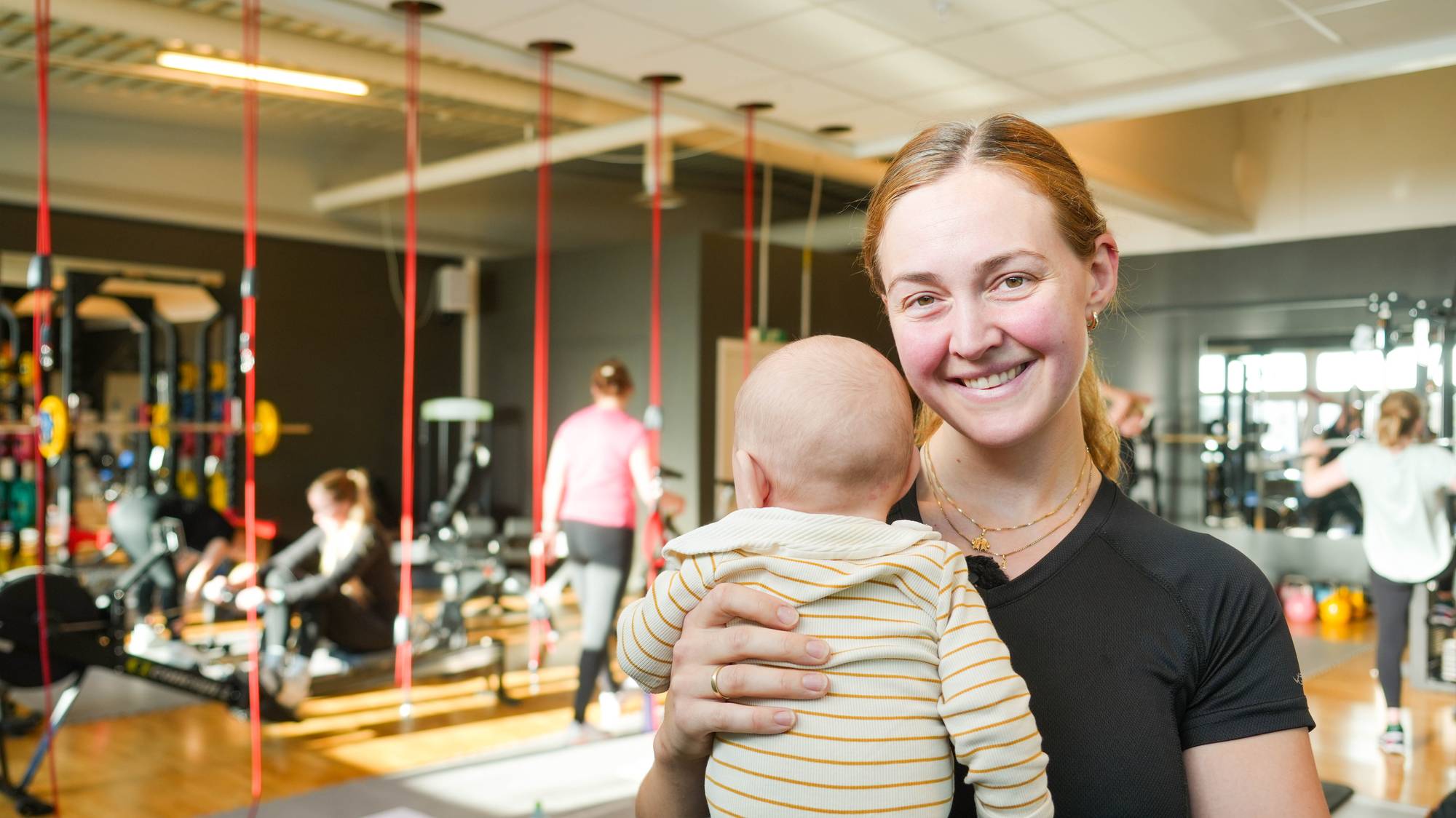 En dame med rødlig hår og sort treningstøy holder en baby på et treningssenter med flere trenende mødre i bakgrunn.