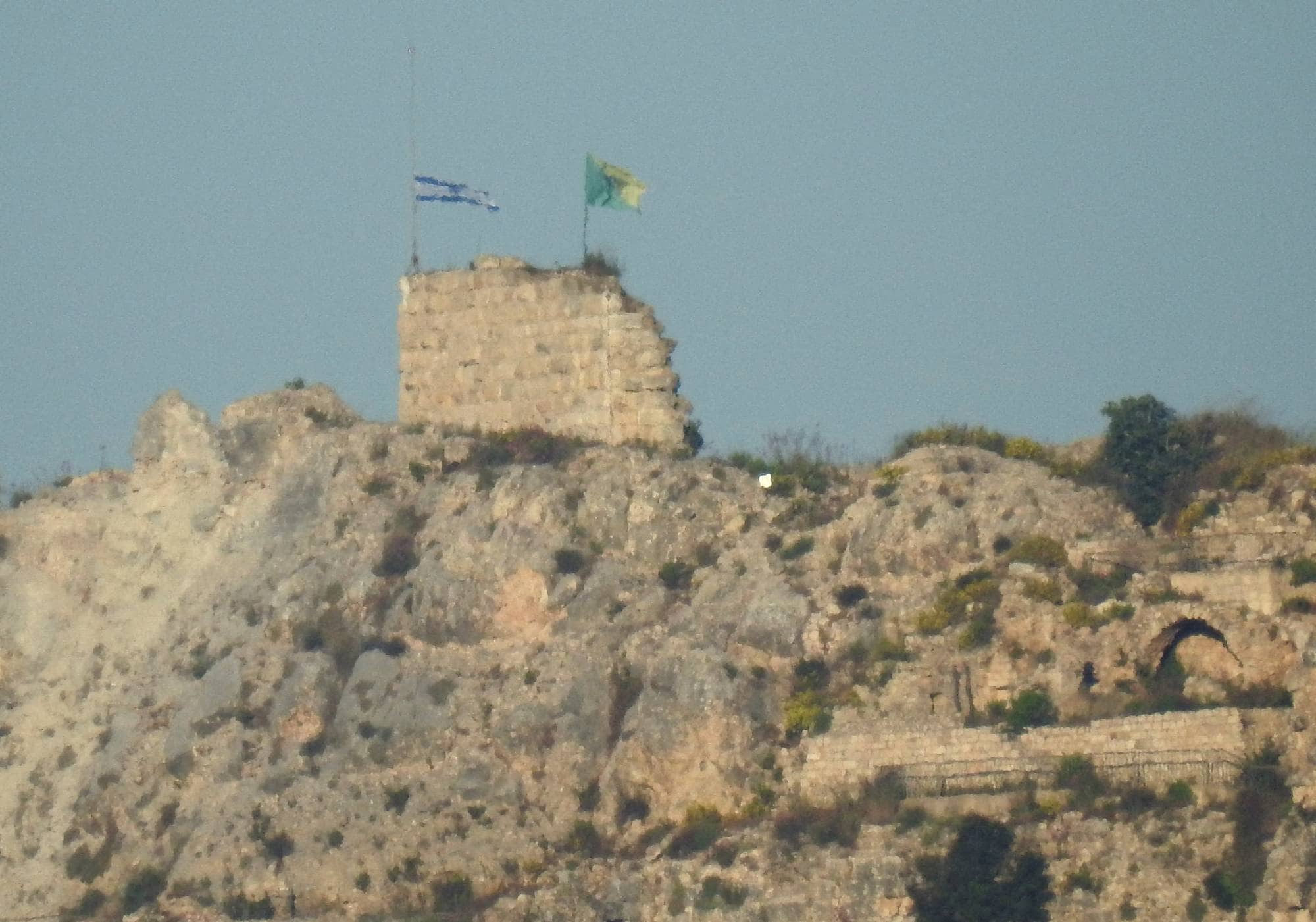Et gammelt fort eller ruin står på en klippe.
To flagg, et med Israels blå davidsstjerne på hvit bakgrunn og et gult flagg, vaier fra toppen av fortet.
Klippen er dekket av steiner og vegetasjon.
Under fortet sees synlige ruiner av steinmurer og buede innganger. (Bildebeskrivelsen er laget av en KI-tjeneste)