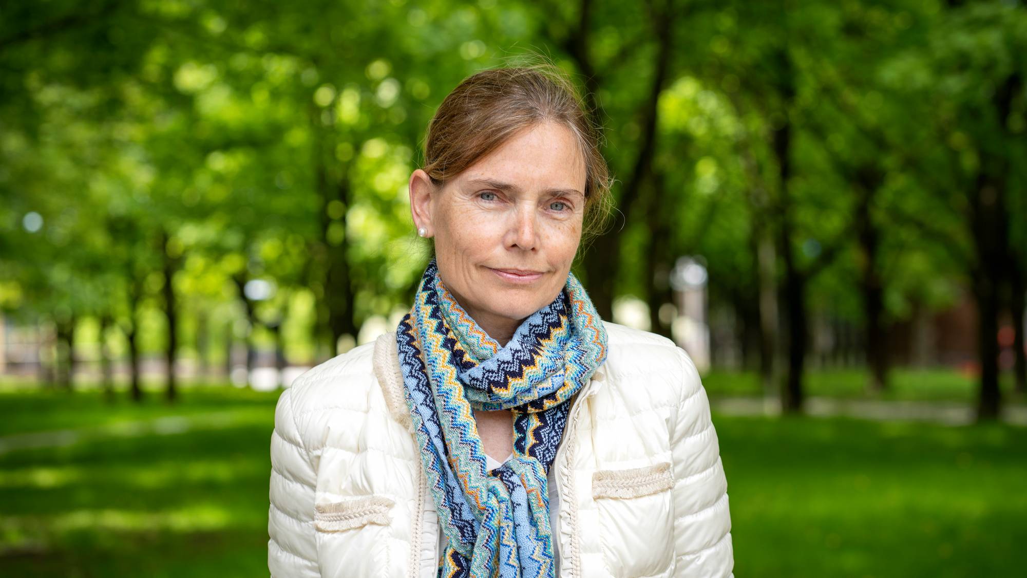Portrett av kvinne ute. Ikledd en hvit tynn boblejakke og et fargerrikt blått og gult skjerf. 