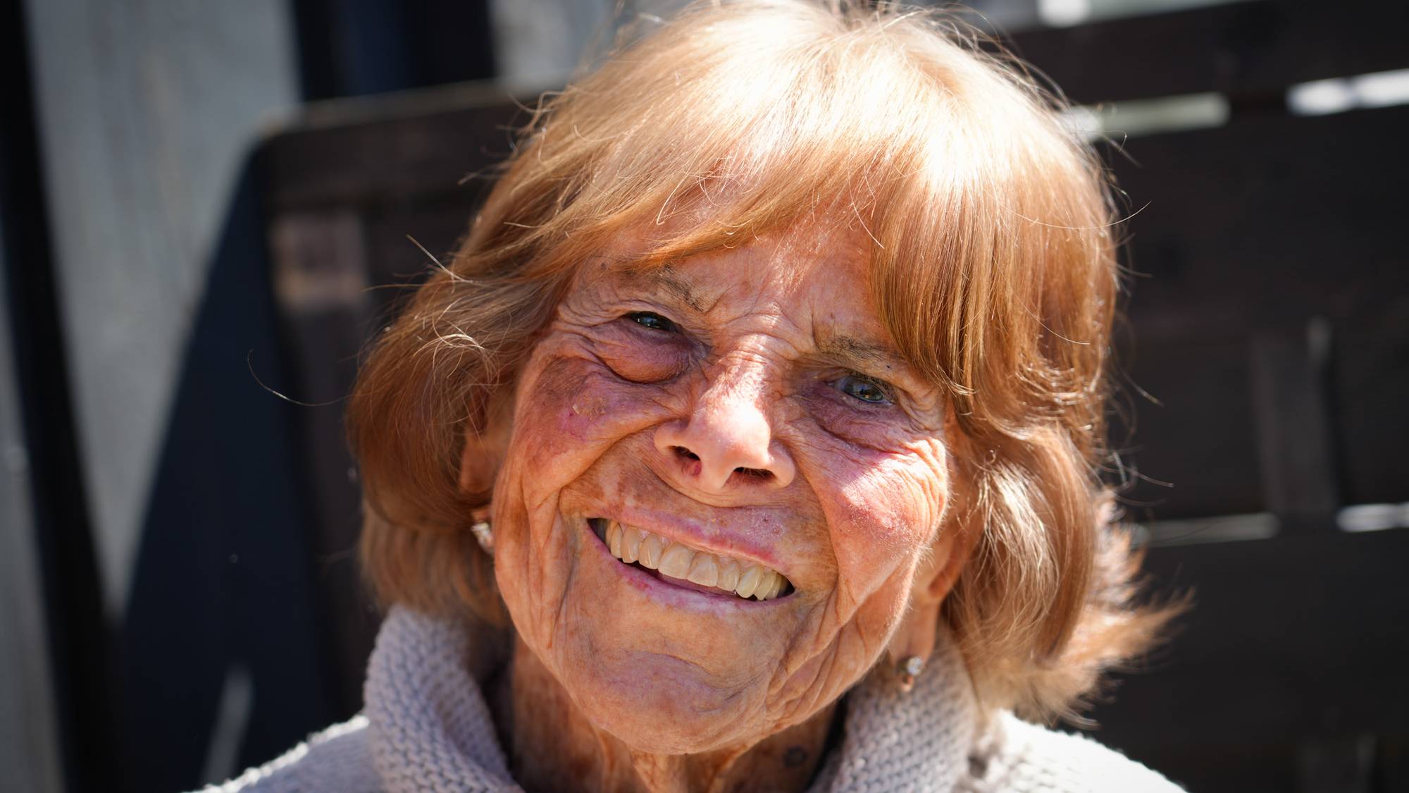 Close-up portrait of an elderly woman smiling. She has fair skin and light brown hair. She is wearing a light-colored knitted sweater. The background is blurred and appears to be outdoors. (Bildebeskrivelsen er laget av en KI-tjeneste)