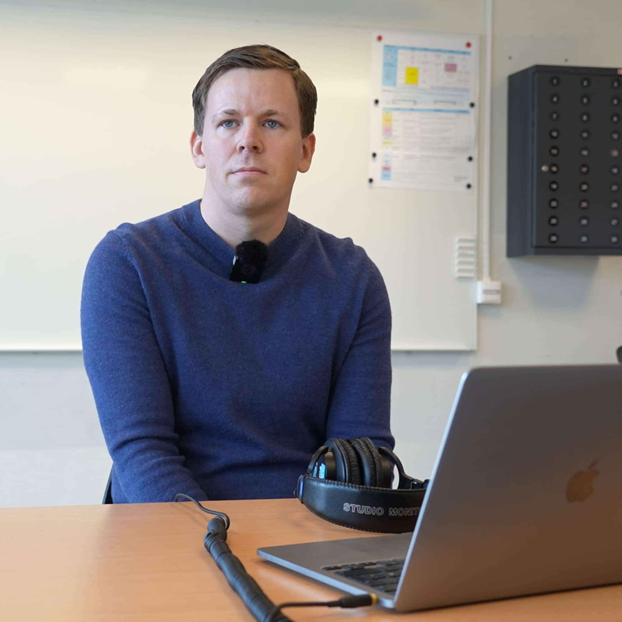 Mann i blå genser sitter foran en bærbar PC i et klasserom med en hvit tavle i bakgrunnen. (Bildebeskrivelsen er laget av en KI-tjeneste)