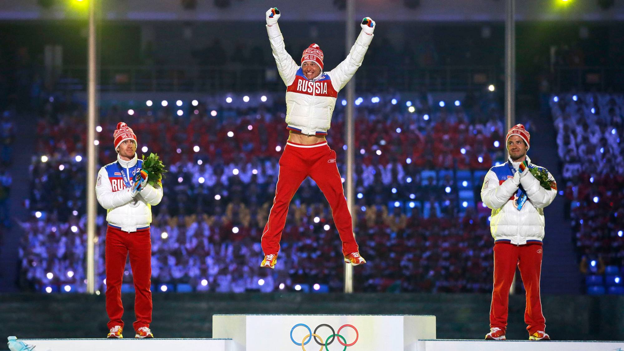 Tre medaljevinnere står på et podium. En av dem hopper i luften med armene hevet. De bærer alle klær med påskriften "RUSSIA". Det er OL-symboler og "SOCHI 2014" på podiet. (Bildebeskrivelsen er laget av en KI-tjeneste)