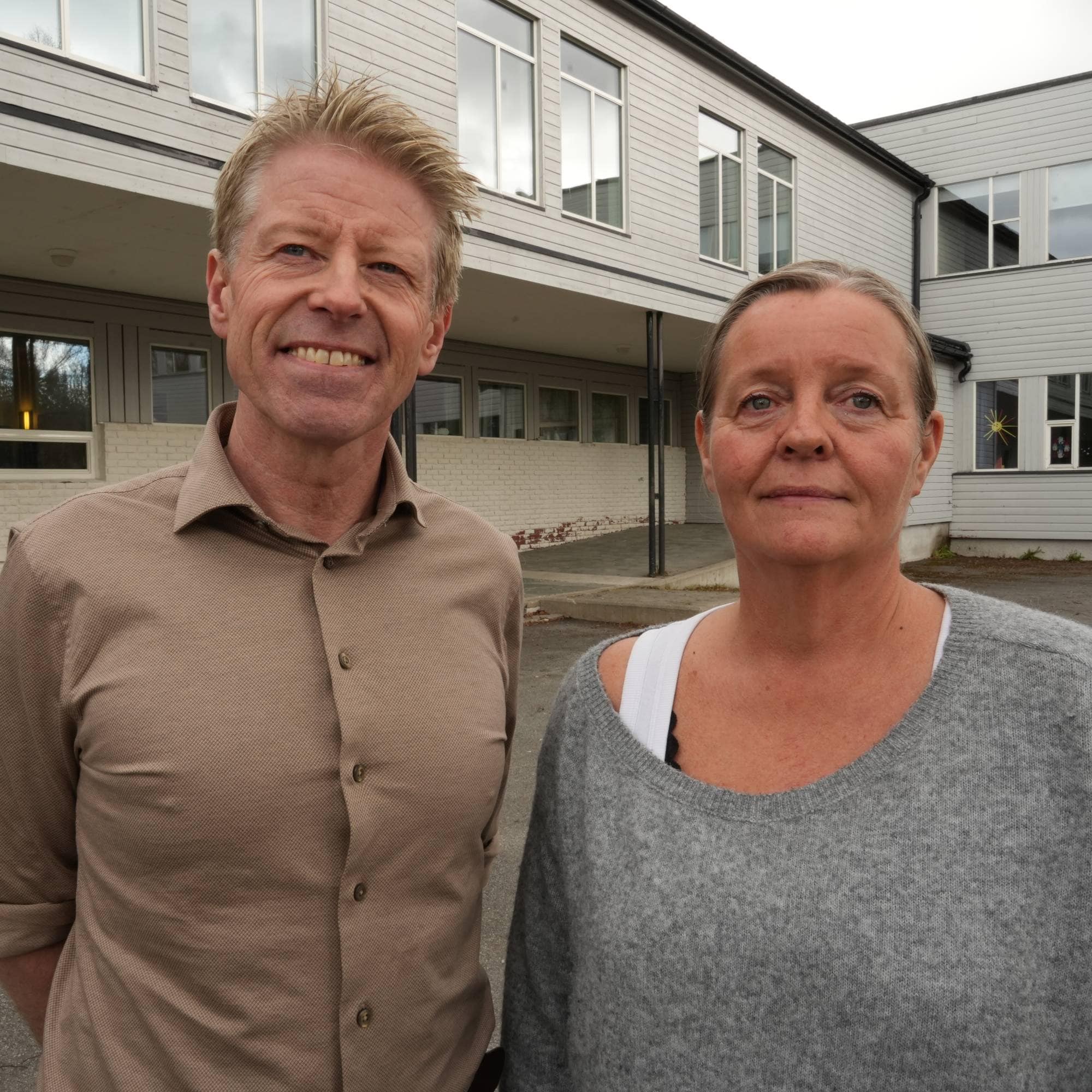 En mann og en dame står ute foran et lyseblått skolebygg. De ser mot kamera. 