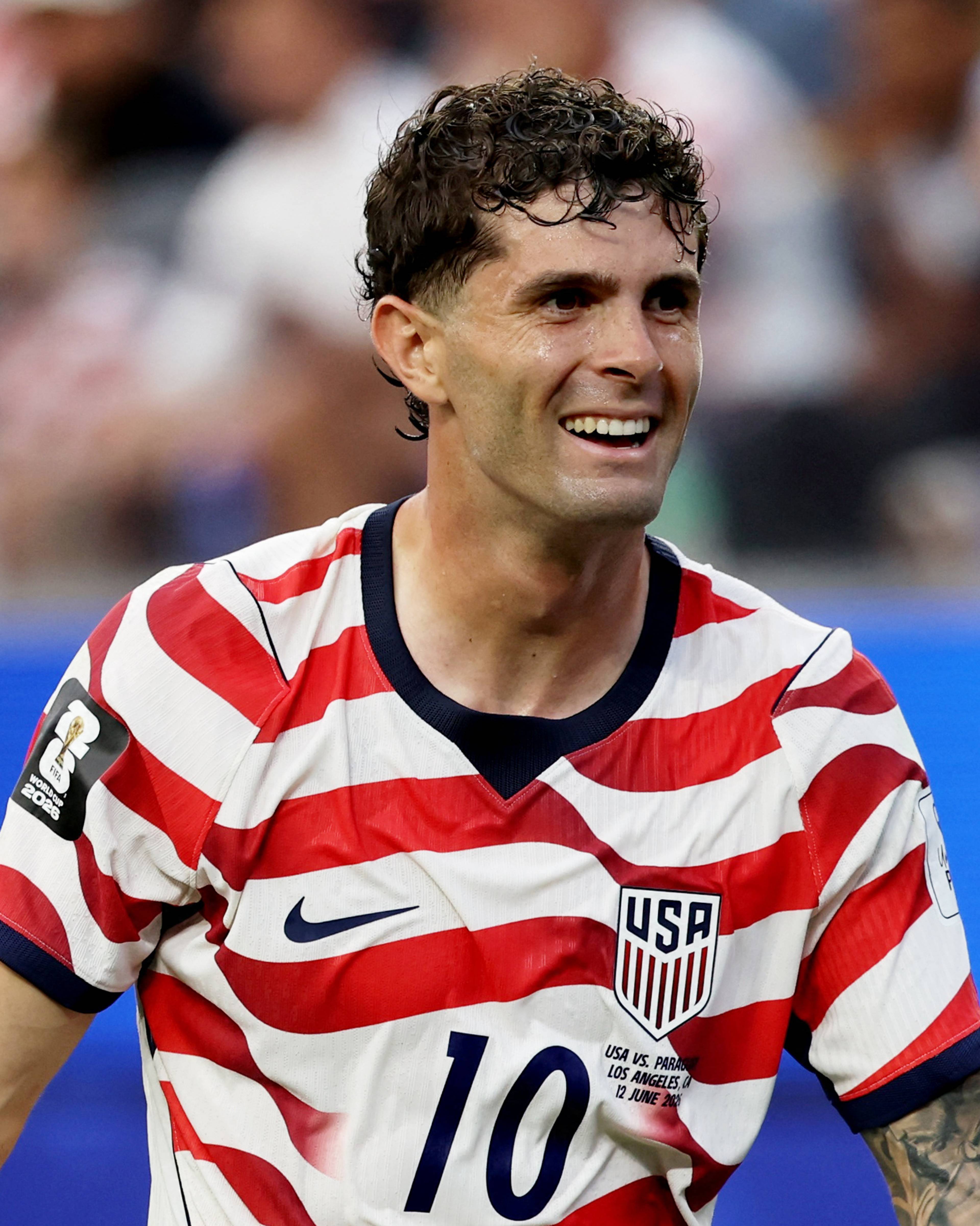 En fotballspiller i USA-drakt med nummer 10, Christian Pulisic, smiler og ser ut mot siden. Han har armene utstrakt. Bak ham er tribuner med publikum i det som ser ut som en fotballarena. (Bildebeskrivelsen er laget av en KI-tjeneste)