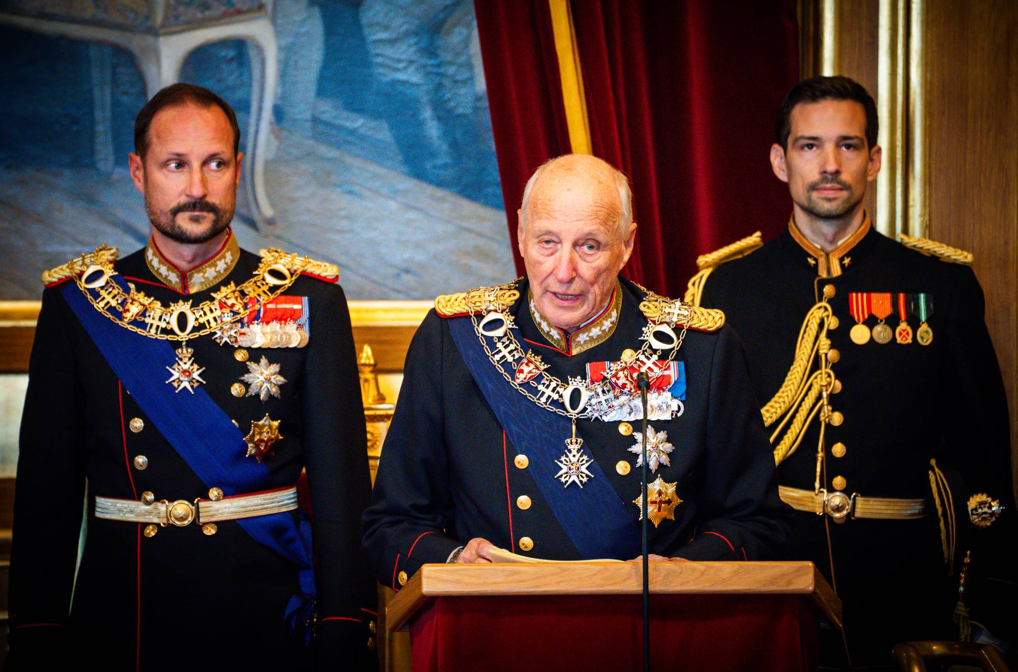 Kong Harald taler fra en talerstol flankert av kronprins Haakon og en adjutant i gallauniform. (Bildebeskrivelsen er laget av en KI-tjeneste)