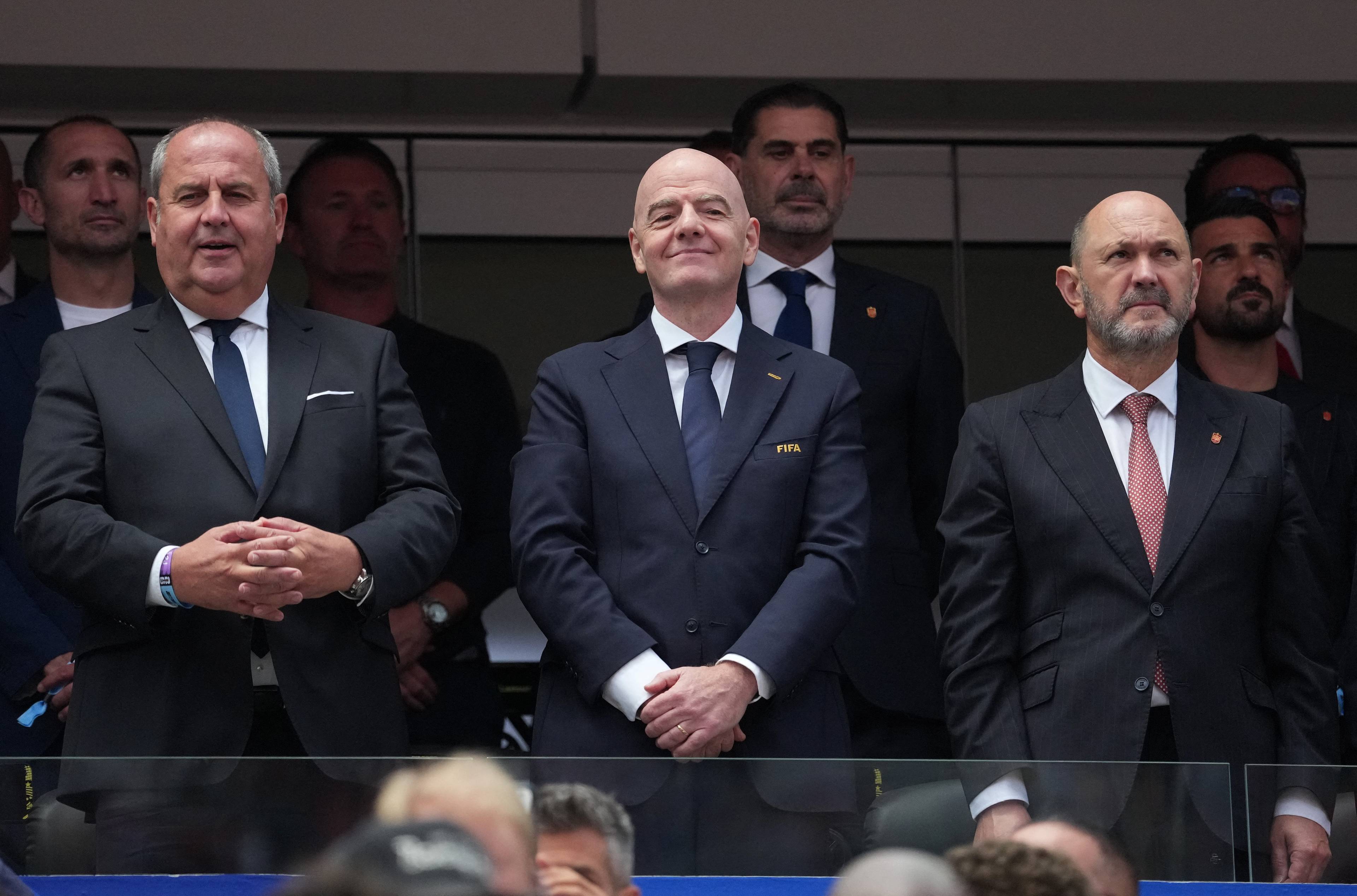 FIFA-president Gianni Infantino flankert av menn i dress på tribunen under ein fotballkamp. (Bildebeskrivelsen er laget av ein KI-teneste)