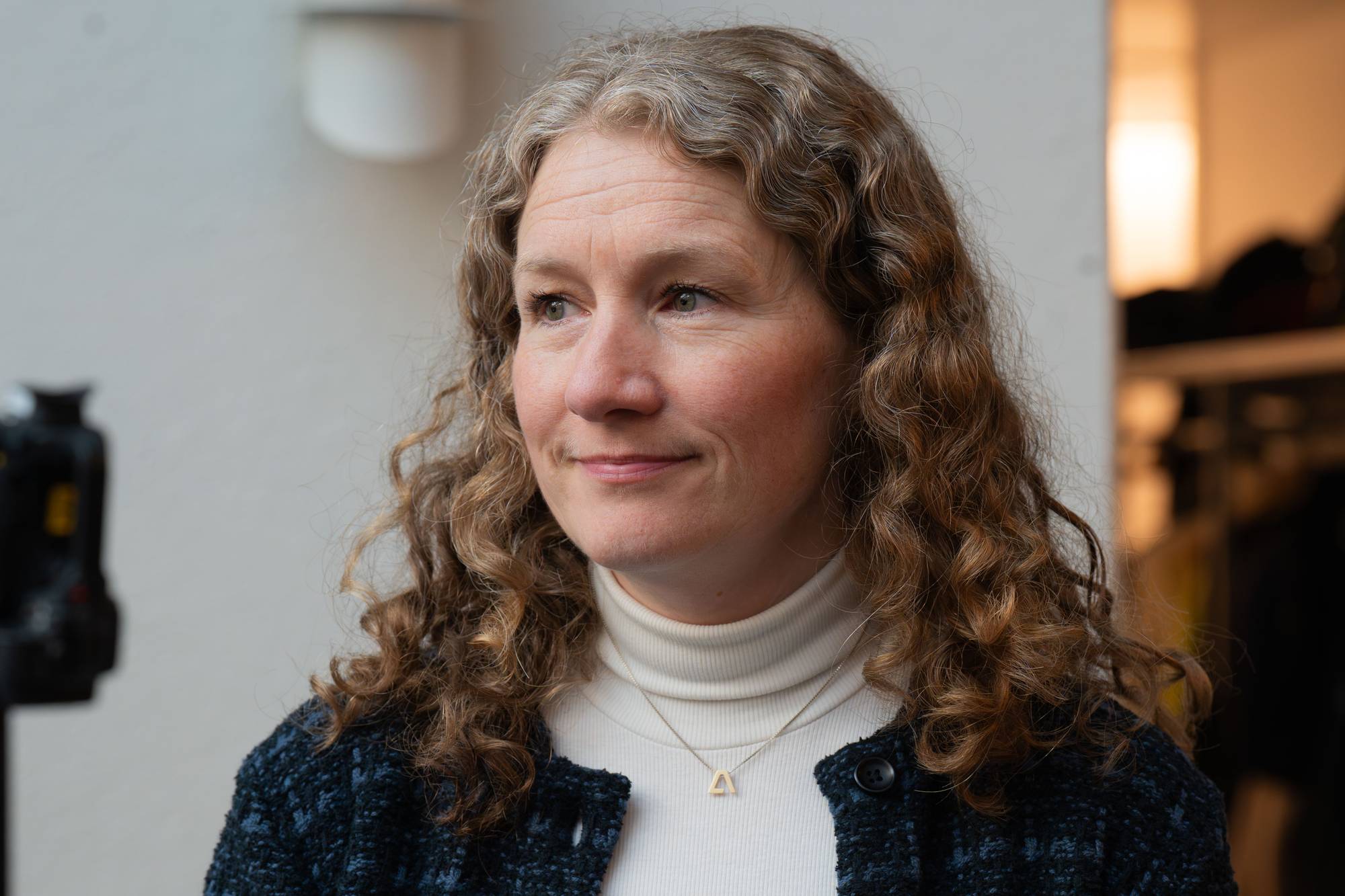 En kvinne med krøllete, lyst hår og en hvit turtleneck ser til siden. Hun har på seg en mørkeblå jakke og et anheng i gull. Til venstre i bildet, i bakgrunnen, er en mikrofon på stativ. Lyssettingen er myk og bakgrunnen er uskarp. (Bildebeskrivelsen er laget av en KI-tjeneste)