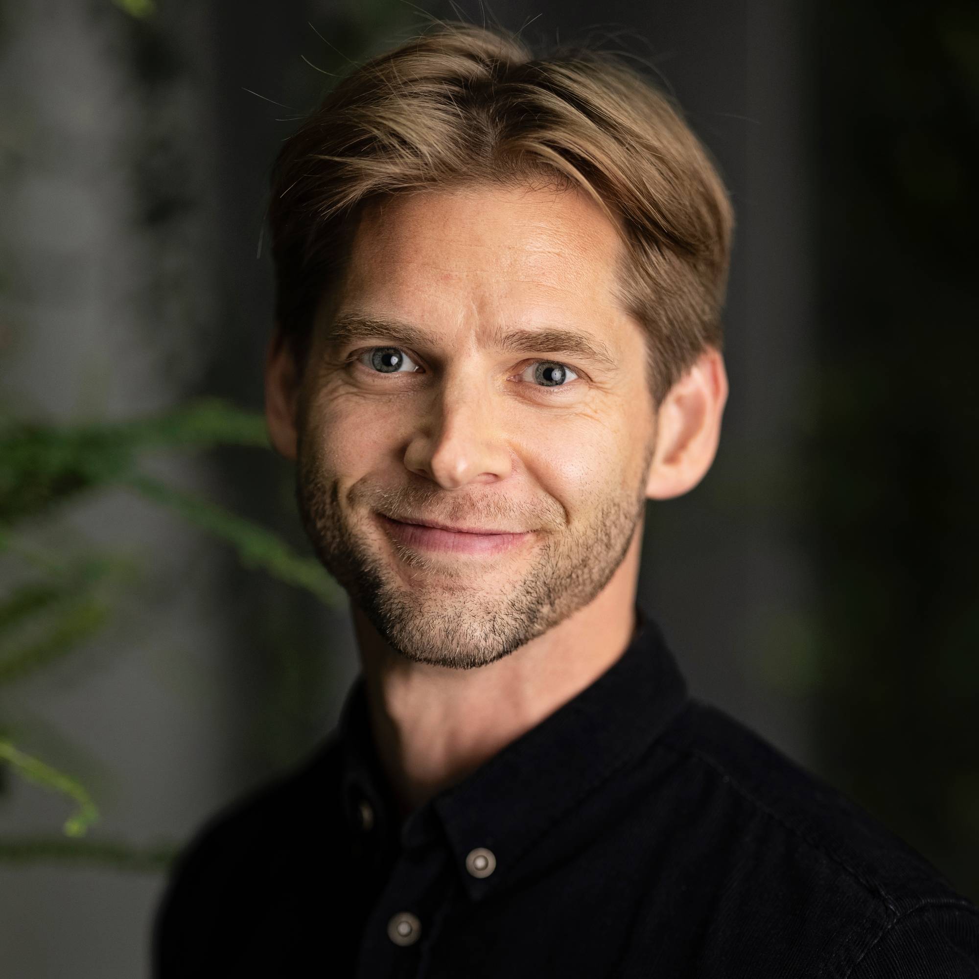 Et portrettbilde av en mann i svart skjorte. Han har brunt hår og skjegg. I bakgrunnen er det grønne planter og en mørk vegg. Mannen ser mot kameraet og smiler. (Bildebeskrivelsen er laget av en KI-tjeneste)