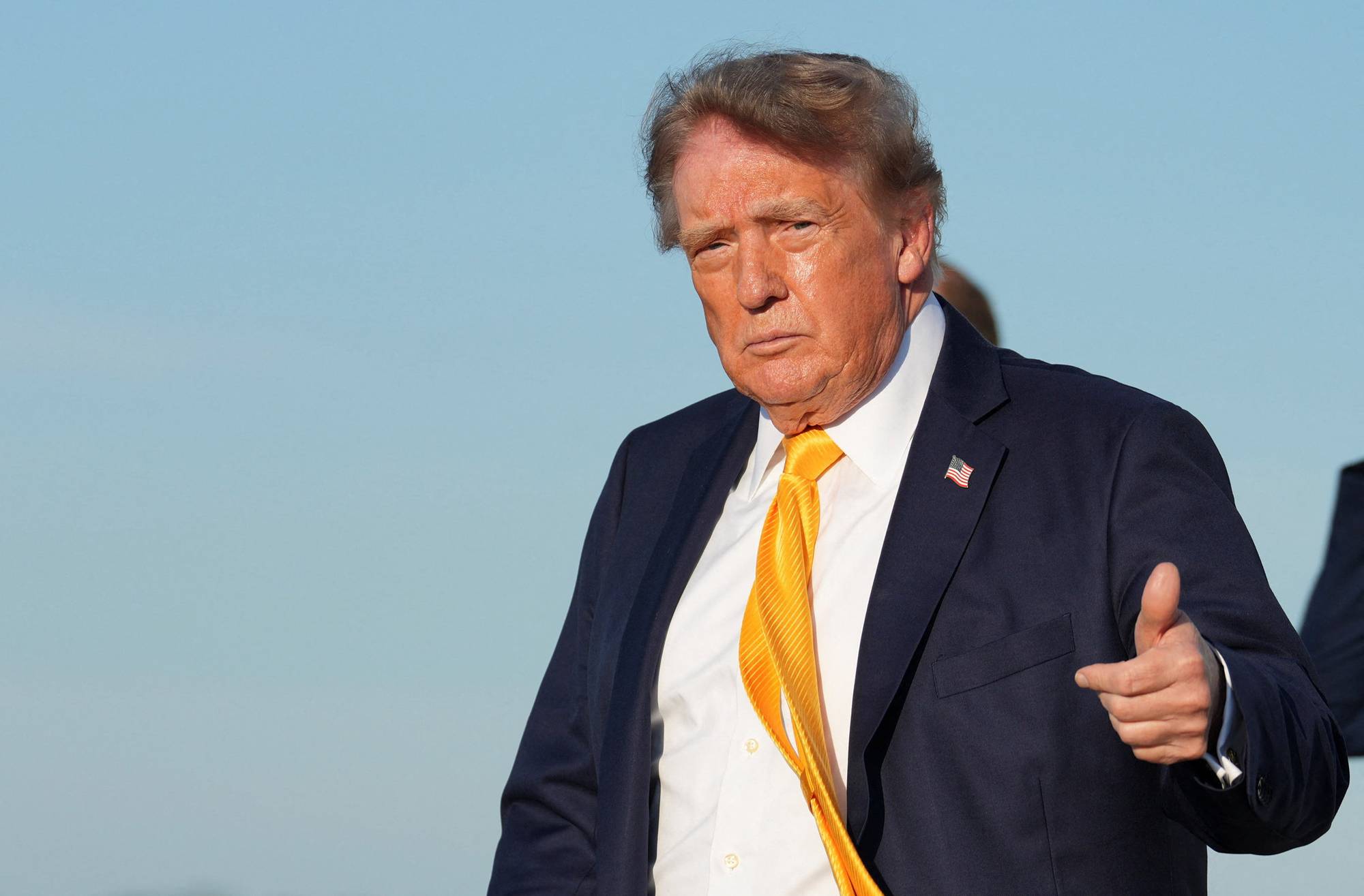 Donald Trump i mørk dress og gult slips viser tommel opp foran en blå himmel. (Bildebeskrivelsen er laget av en KI-tjeneste)