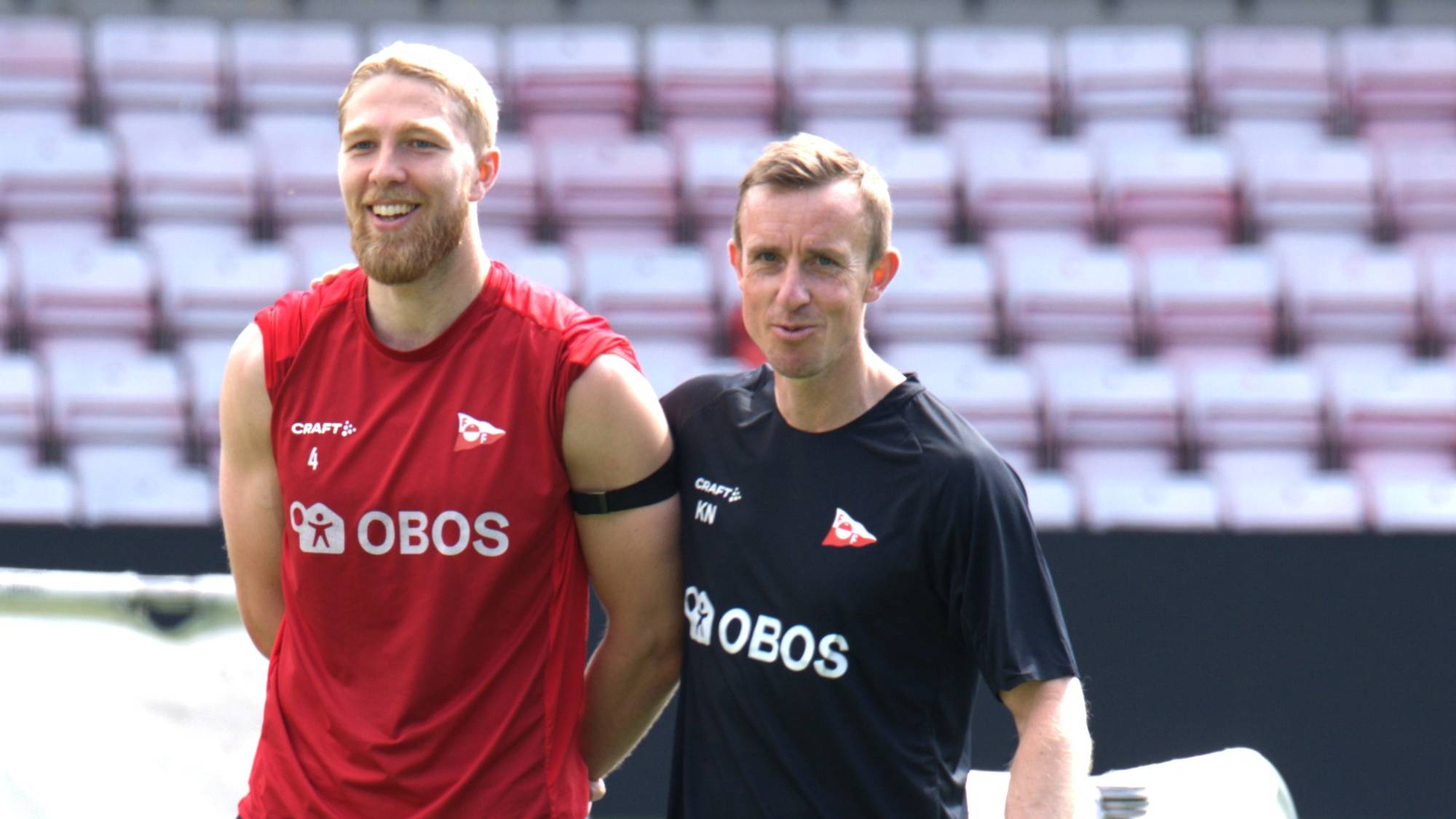 To menn smiler på et kunstgress og holder rundt hverandre, den ene med rød singlet, den andre med svart t-skjorte.