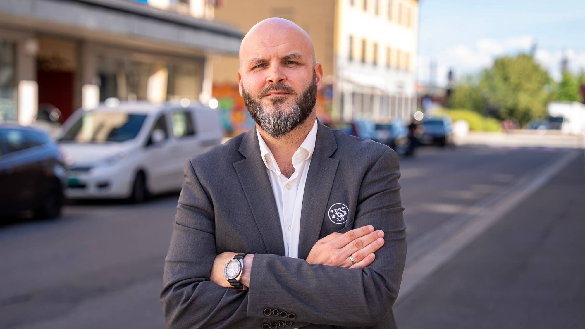 Skallet mann med skjegg i dress står med armene i kors i en bygate. (Bildebeskrivelsen er laget av en KI-tjeneste)
