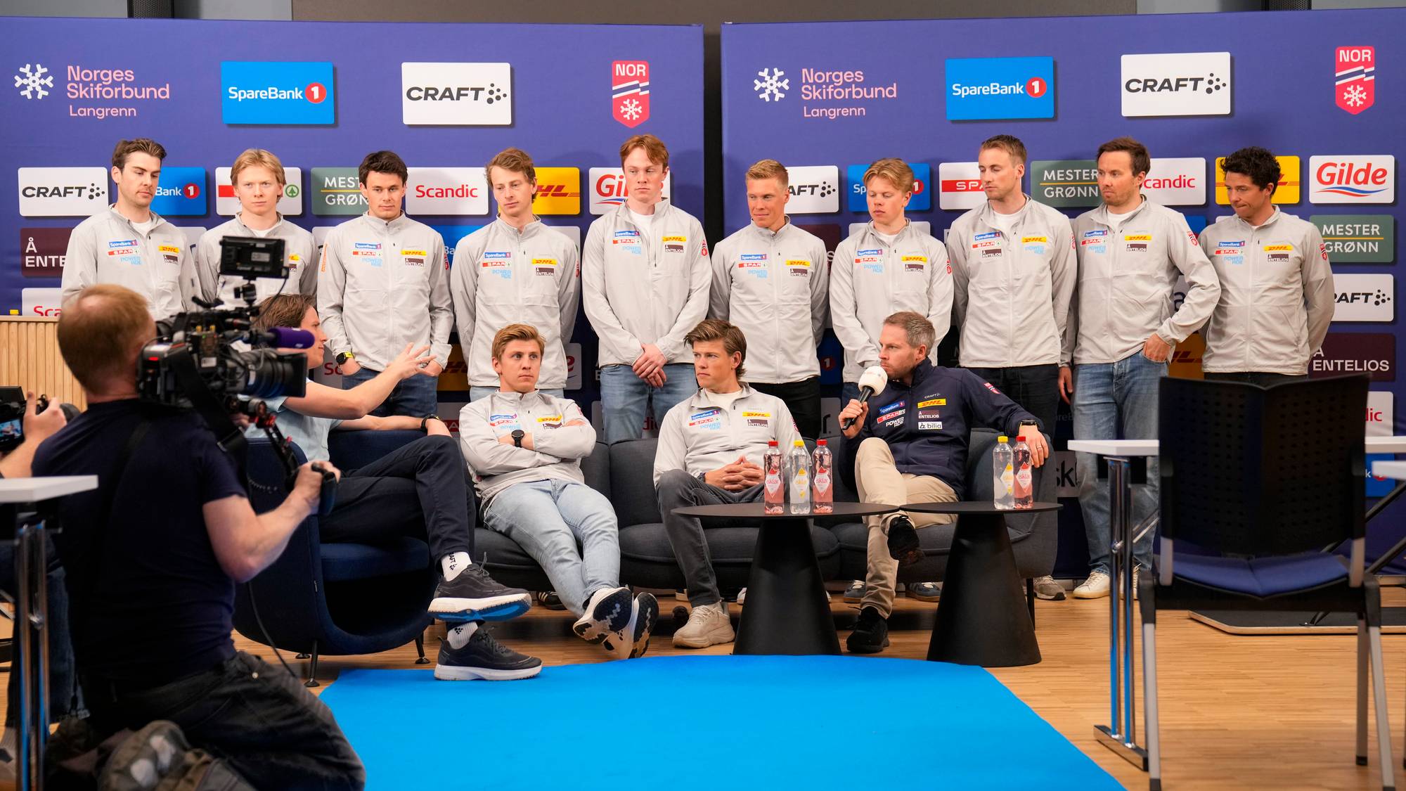 En gruppe menn står samlet foran en pressevegg. Én person sitter på en sofa og holder en mikrofon. En annen person sitter på en stol og filmer. Bak mennene er det en blå bakgrunn med sponsorlogoer. (Bildebeskrivelsen er laget av en KI-tjeneste)