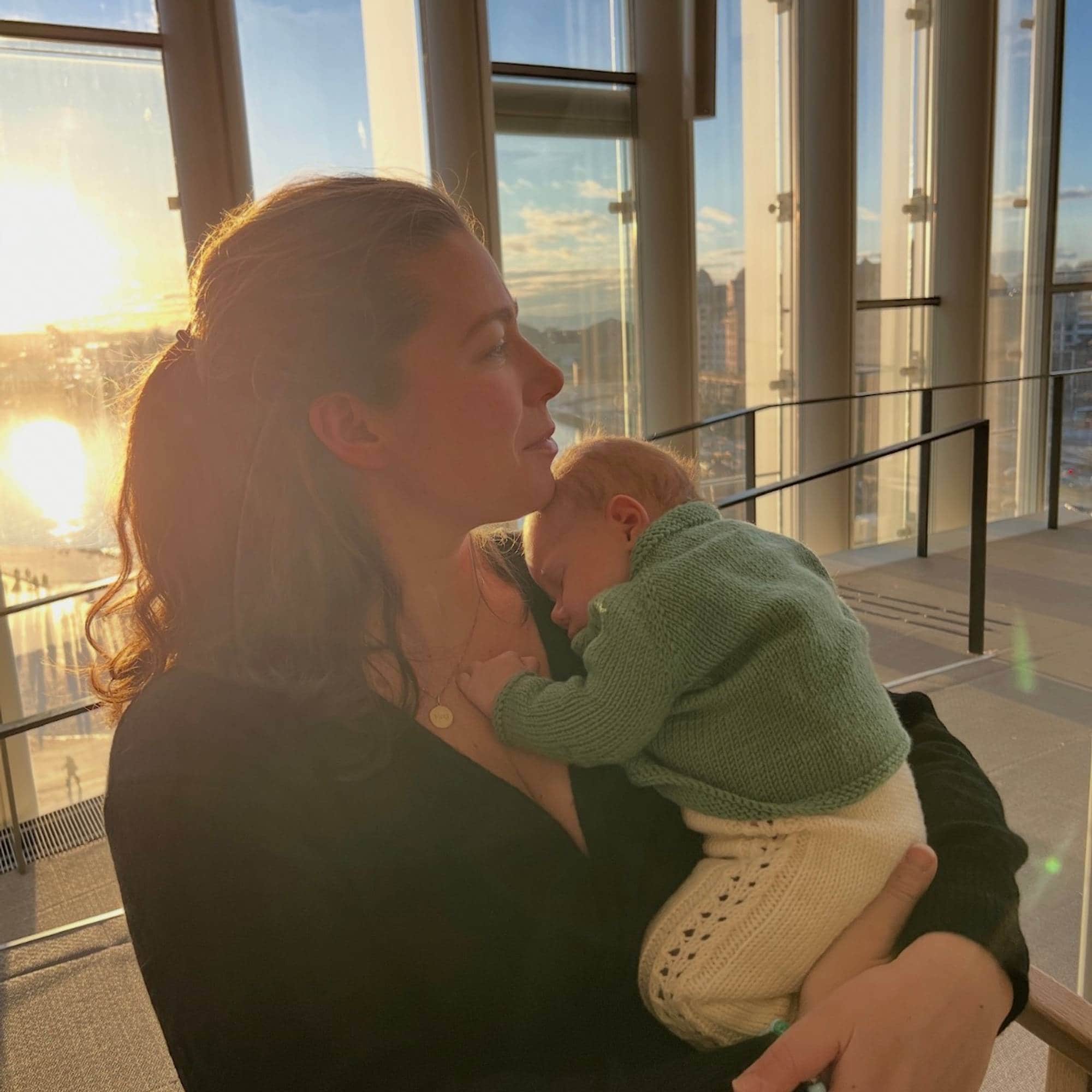Kvinne holder et sovende spedbarn foran et vindu med solnedgang i bakgrunnen.