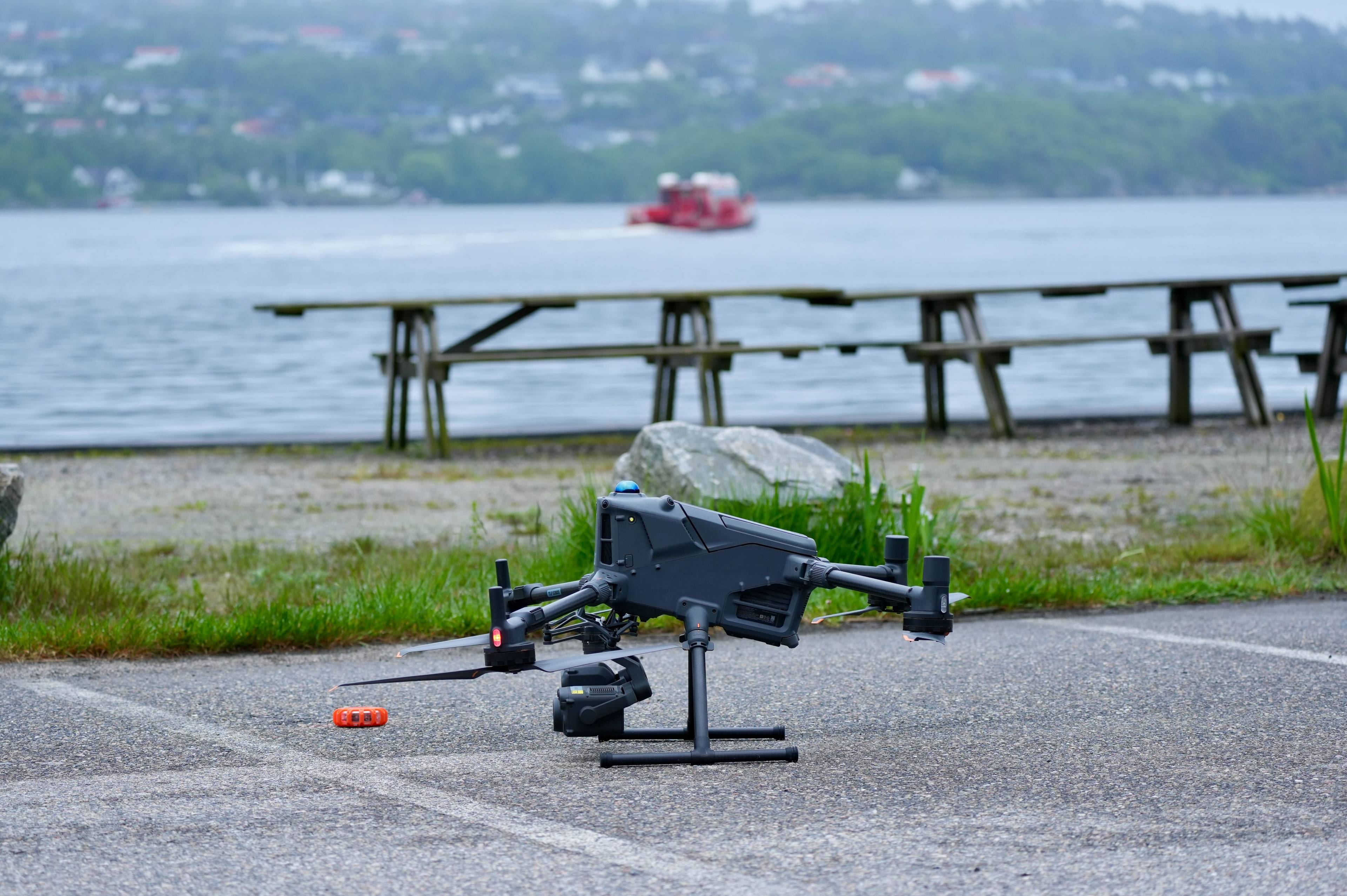 Et mørkt drone står på et stativ på et asfaltert område. En rød båt sees i det fjerne på vannet. En rekke benker langs vannkanten er synlige bak dronen. (Bildebeskrivelsen er laget av en KI-tjeneste)