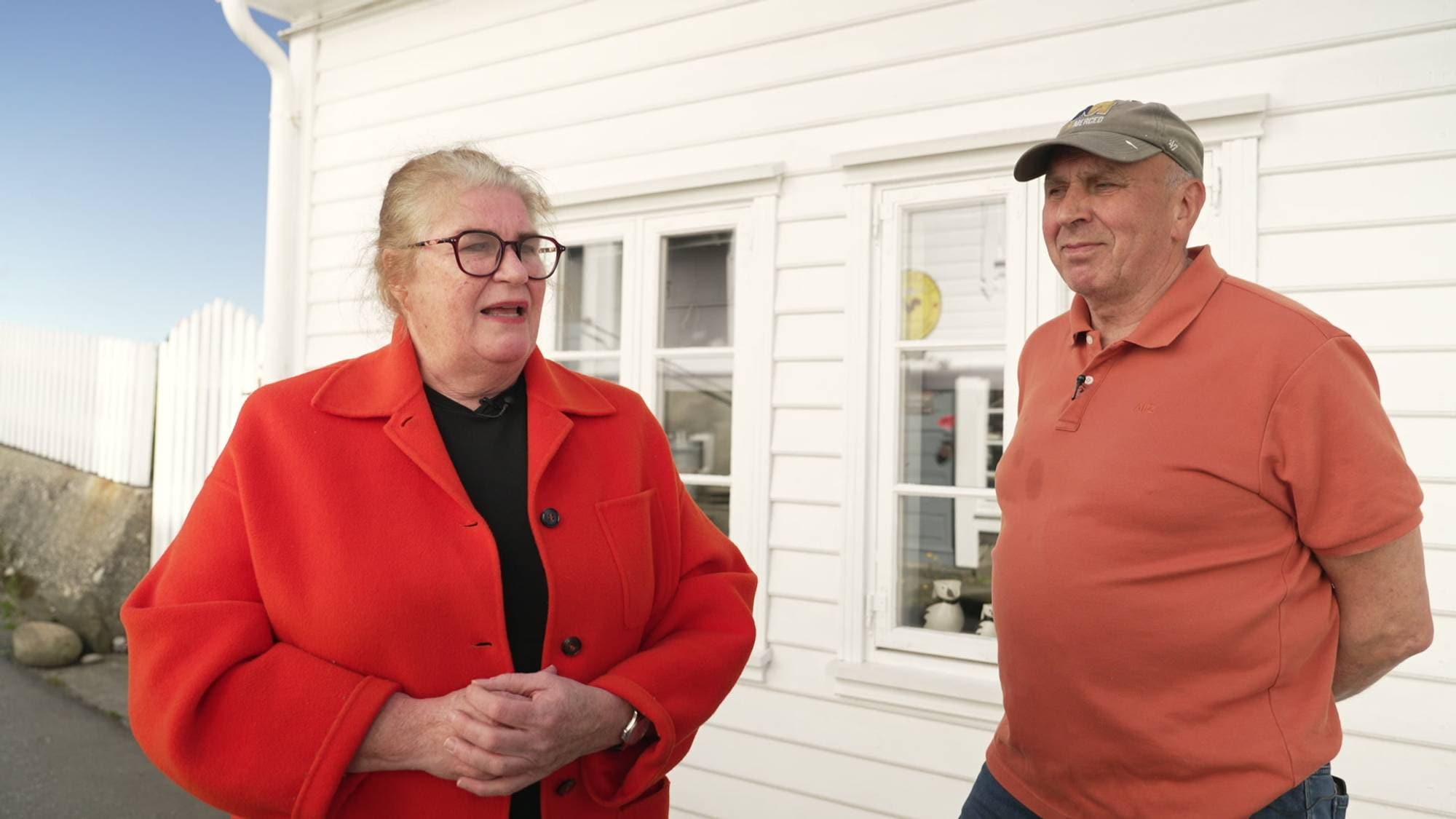 To personer står foran et hvitt hus. Kvinnen til venstre har på seg en oransje jakke og briller. Mannen til høyre har på seg en oransje t-skjorte og en caps. (Bildebeskrivelsen er laget av en KI-tjeneste)