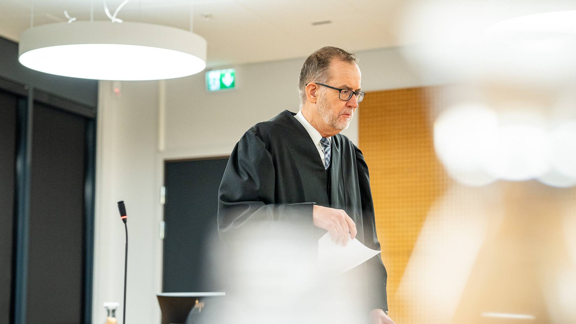 Statsadvokaten holder et papirark. Omgivelsene inkluderer moderne møbler og belysning. En mikrofon er synlig på bordet foran dommeren. Rettssalen ser ut til å være godt opplyst og organisert.