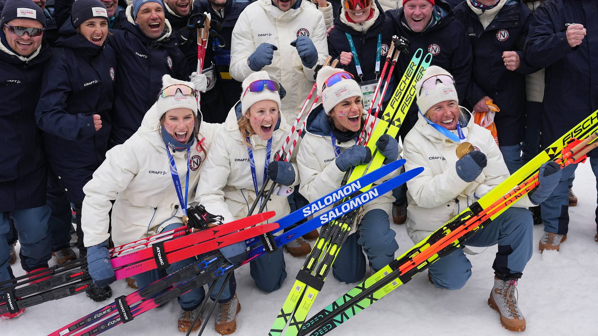 En gruppe skiskyttere feirer en seier med medaljer og ski.
De er kledd i hvite jakker og luer med "NORGE" på.
De holder langrennsski og har medaljer rundt halsen.
Bakgrunnen viser et vinterlandskap med snødekte trær. (Bildebeskrivelsen er laget av en KI-tjeneste)