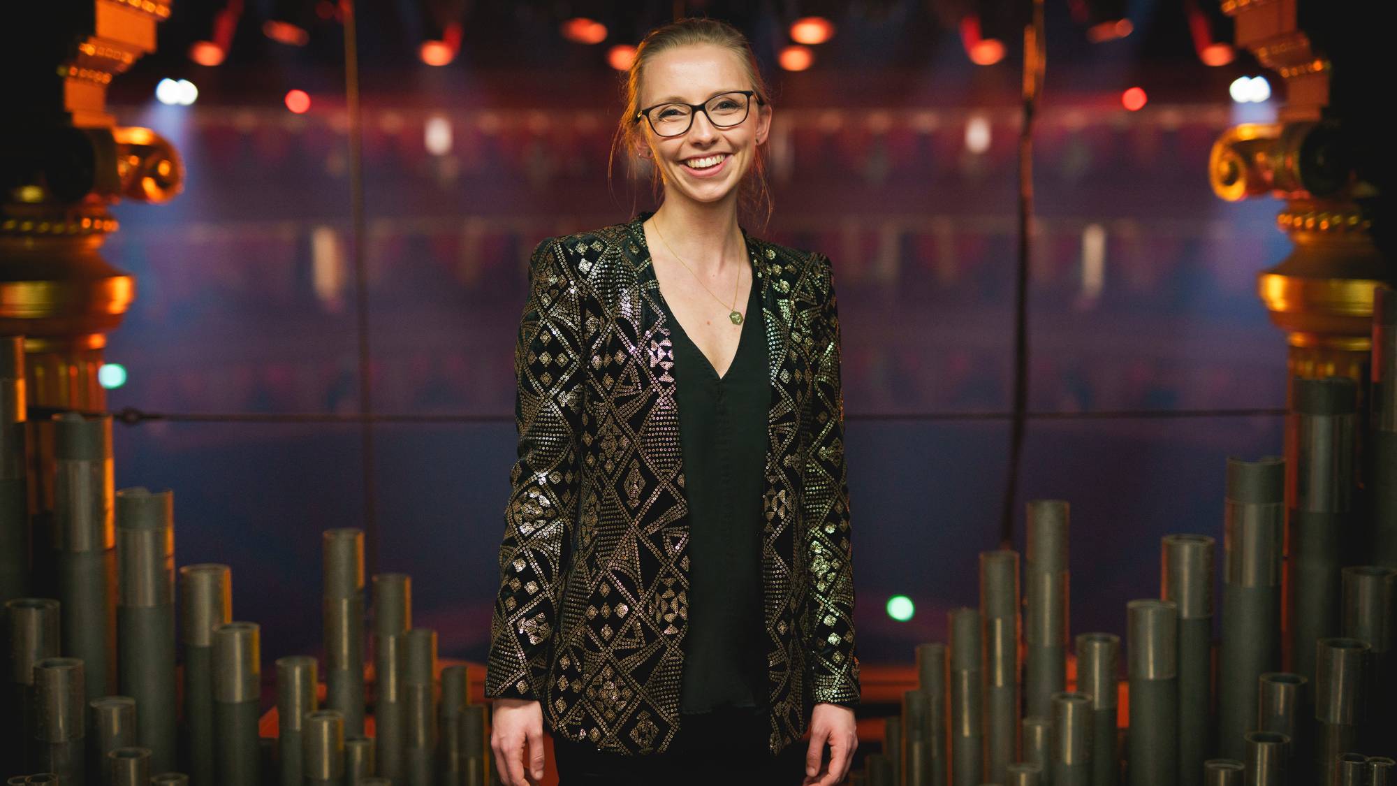 Bildet viser organisten Anna Lapwood i svart dressjakke med sølvmønster foran lyssatte orgelpiper.