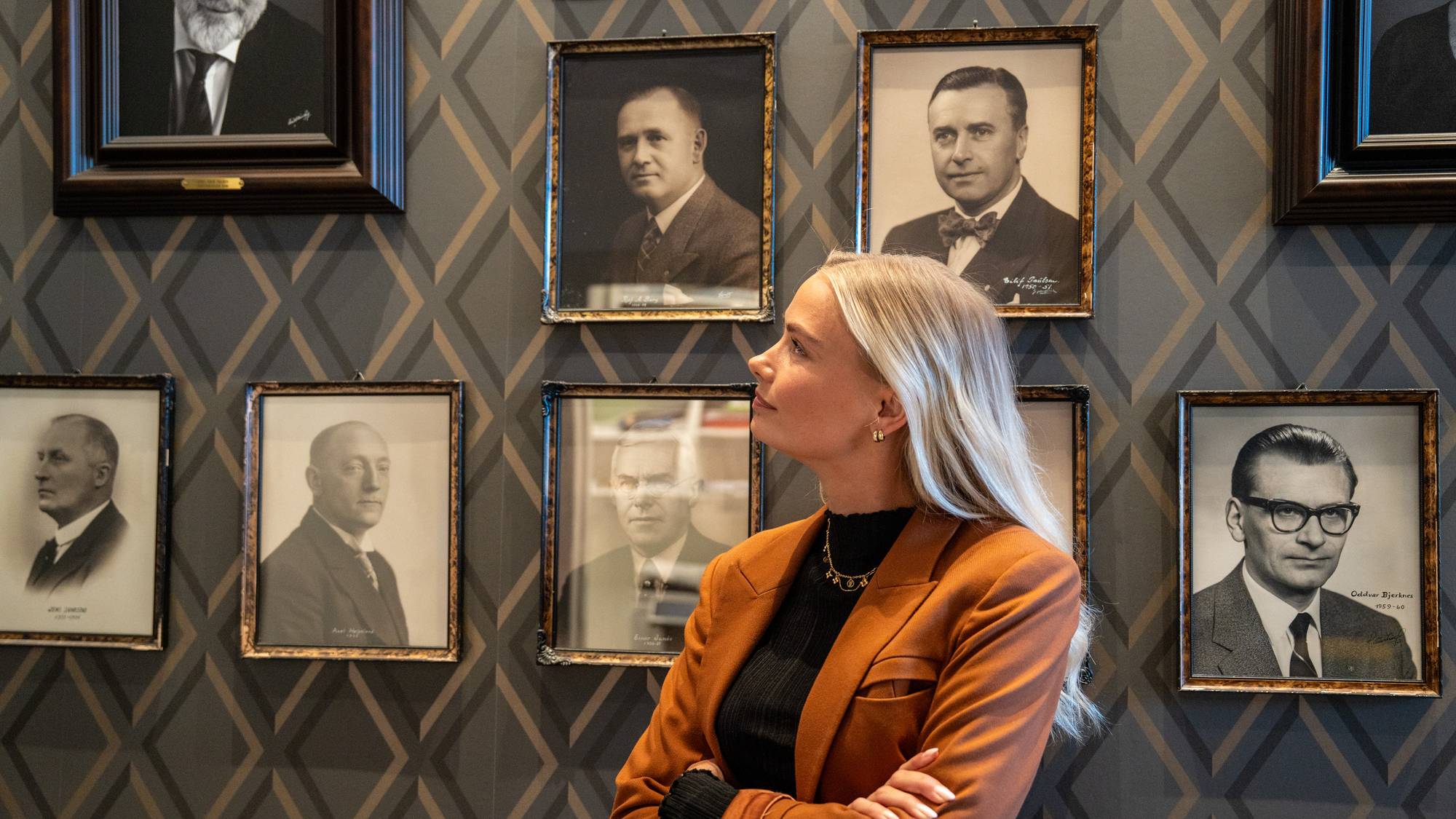 En kvinne med blondt hår står foran en vegg med portrettbilder. Bildene er i ulike størrelser og rammer. Kvinnen er kledd i en brun blazer over en svart genser. 