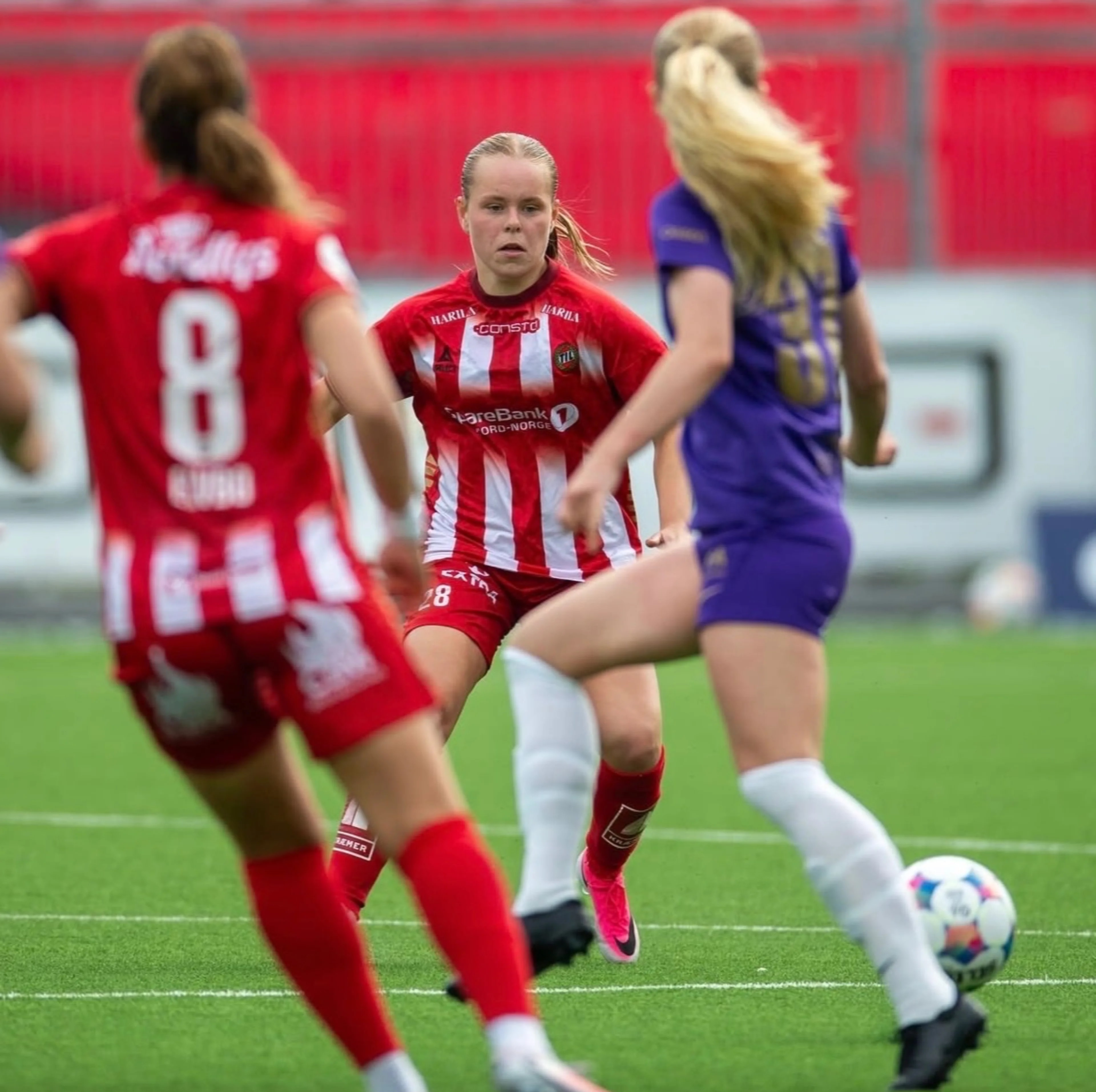 Fotballspillere i røde og lilla drakter kjemper om ballen på en gressbane. (Bildebeskrivelsen er laget av en KI-tjeneste)