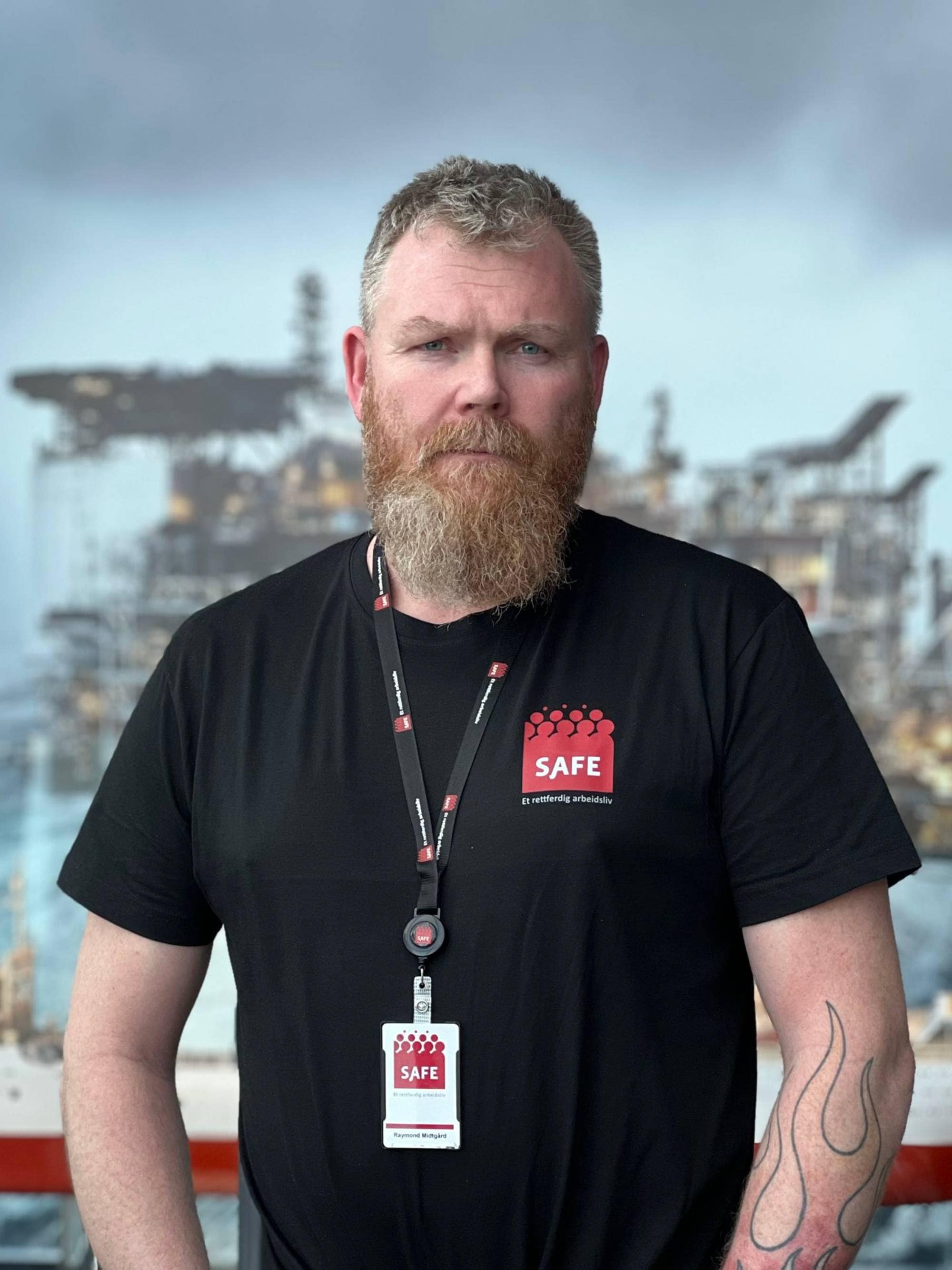Forbundsleder Raymond Midtgård i Safe ser rett i kamera. Han har langt skjegg og kortklipt hår. Han har på seg svart t-skjorte med rød SAFE logo. På armen har han tatovering av flammer. I bakgrunnen ser man en båt og en oljerigg. 