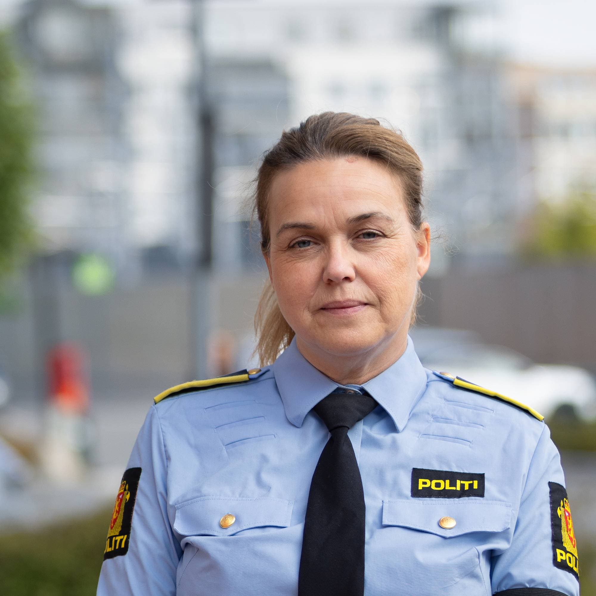 Politikvinne med lyseblå skjorte, mørkt slips og mørke bukser. Håret er festet i en hestehale. 