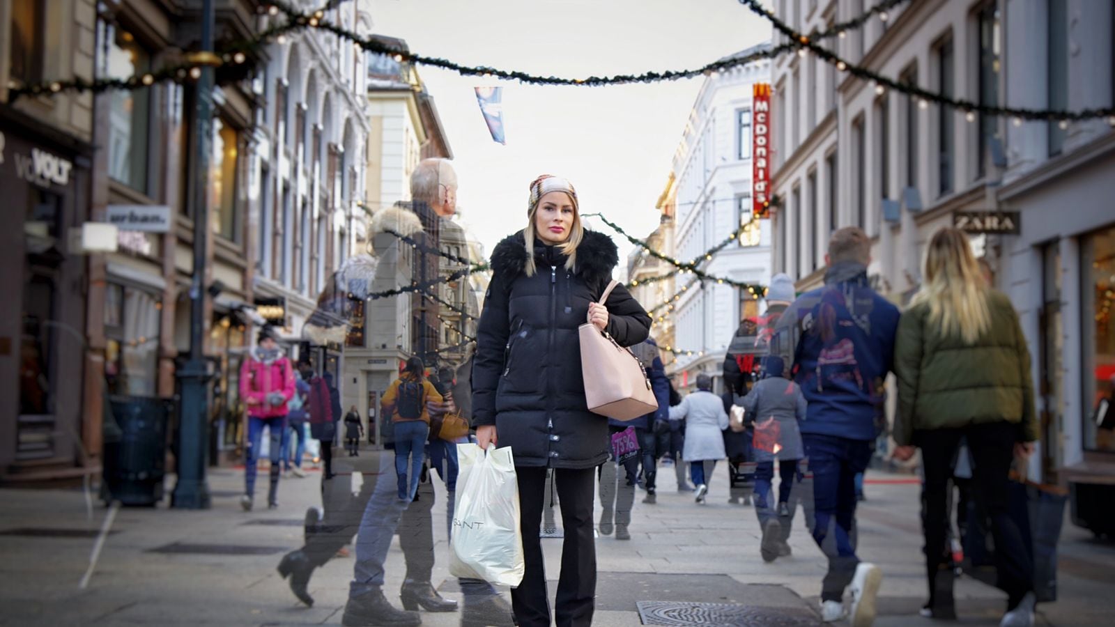 Ung kvinne med handleposer står midt på Karl Johans gate i Oslo og  folk haster forbi henne på begge sider. Vinter.