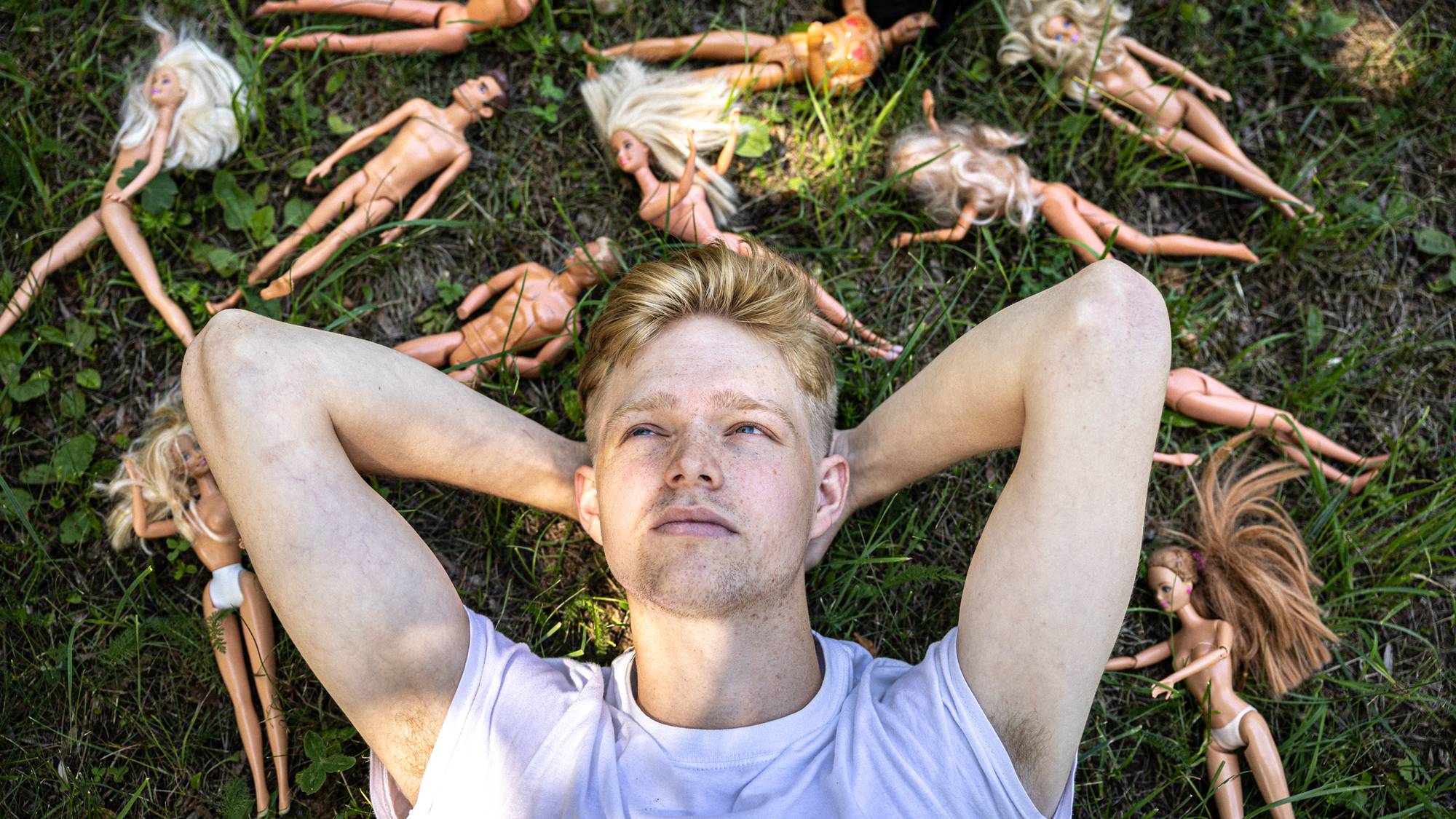 Et portrett av Thomas som ligger med hendene bak hodet på en gressplen. Rundt ham er det strødd barbie-dukker. Thomas ser opp til siden med en undrende tanke i ansiktet. Thomas har rødt kort hår og hvit t-skjorte.