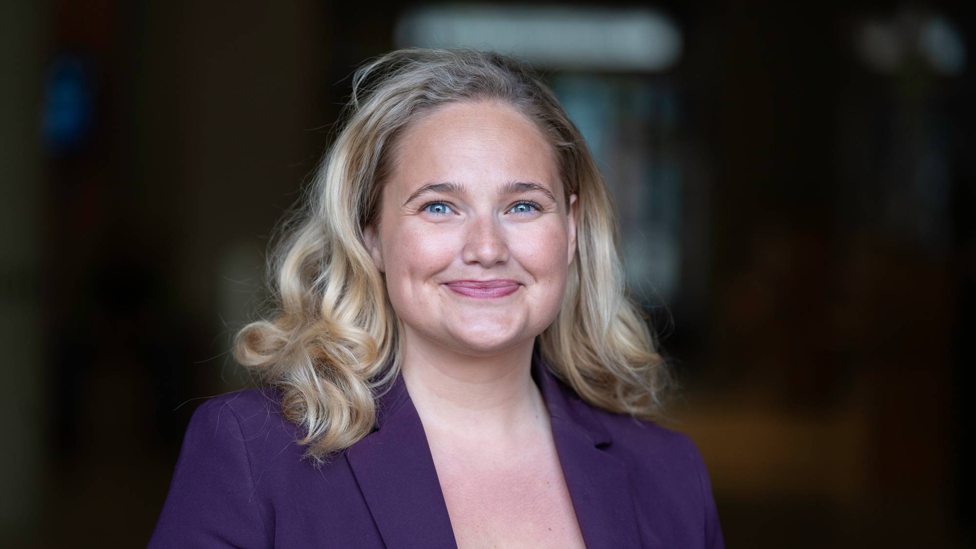 en kvinne med blondt hår og lille dressjakke smiler til kamera