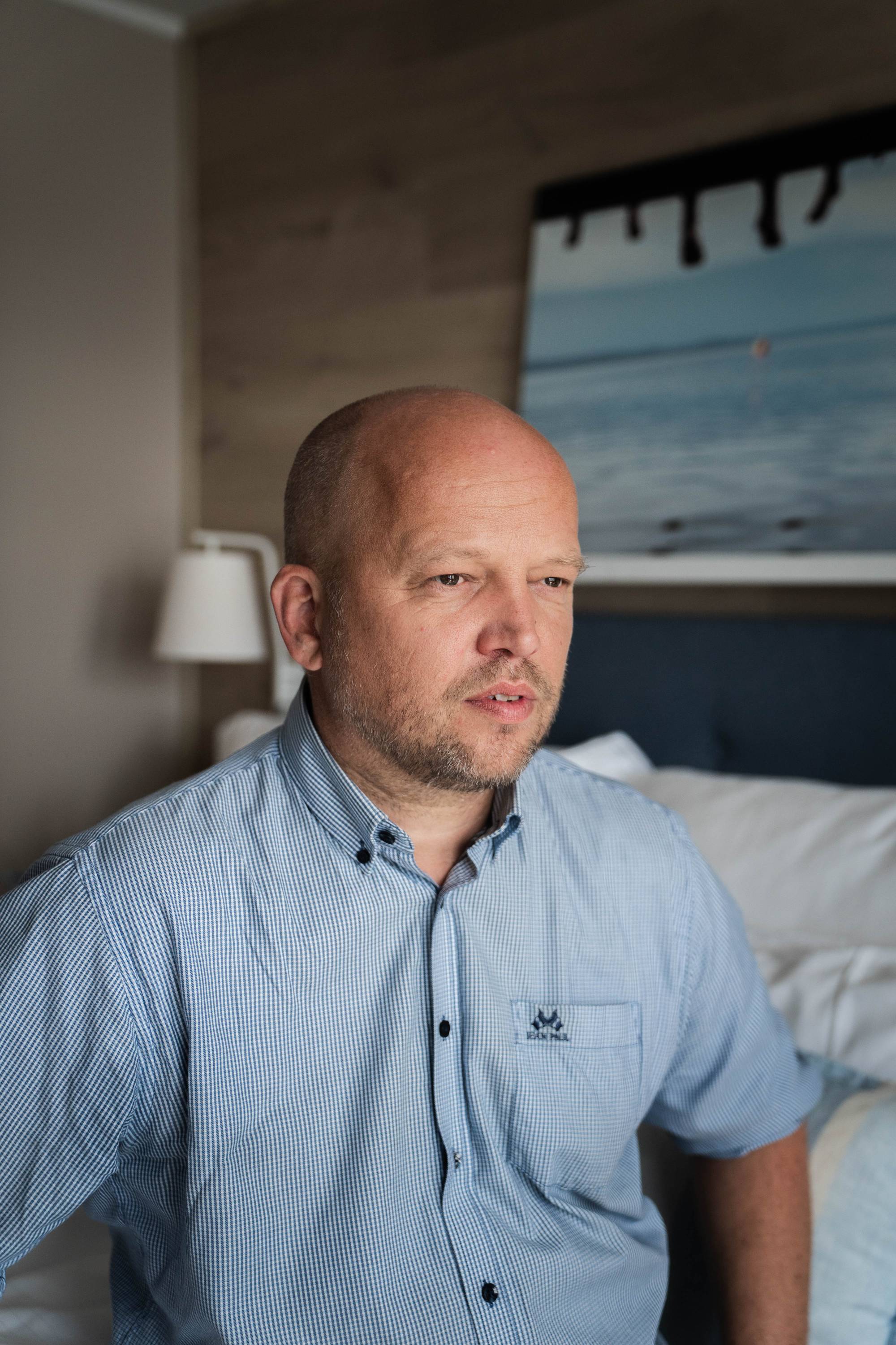 En skallet mann i blå skjorte sitter på et soverom. (Bildebeskrivelsen er laget av en KI-tjeneste)