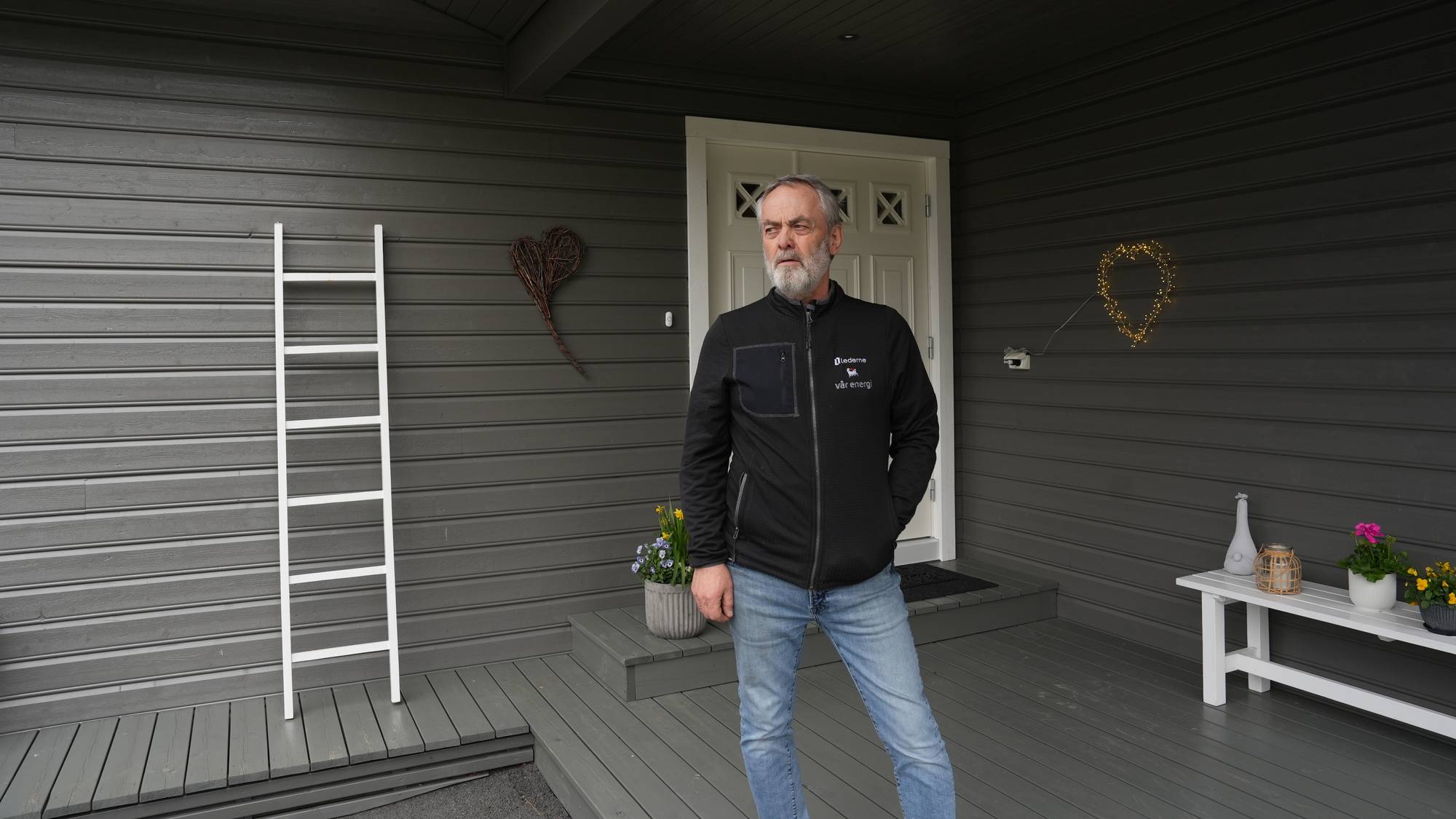 En mann i svart jakke og jeans står foran et hus. Til venstre for mannen står en hvit stige. Til høyre på husveggen henger en hjerteformet lyslenke. (Bildebeskrivelsen er laget av en KI-tjeneste)