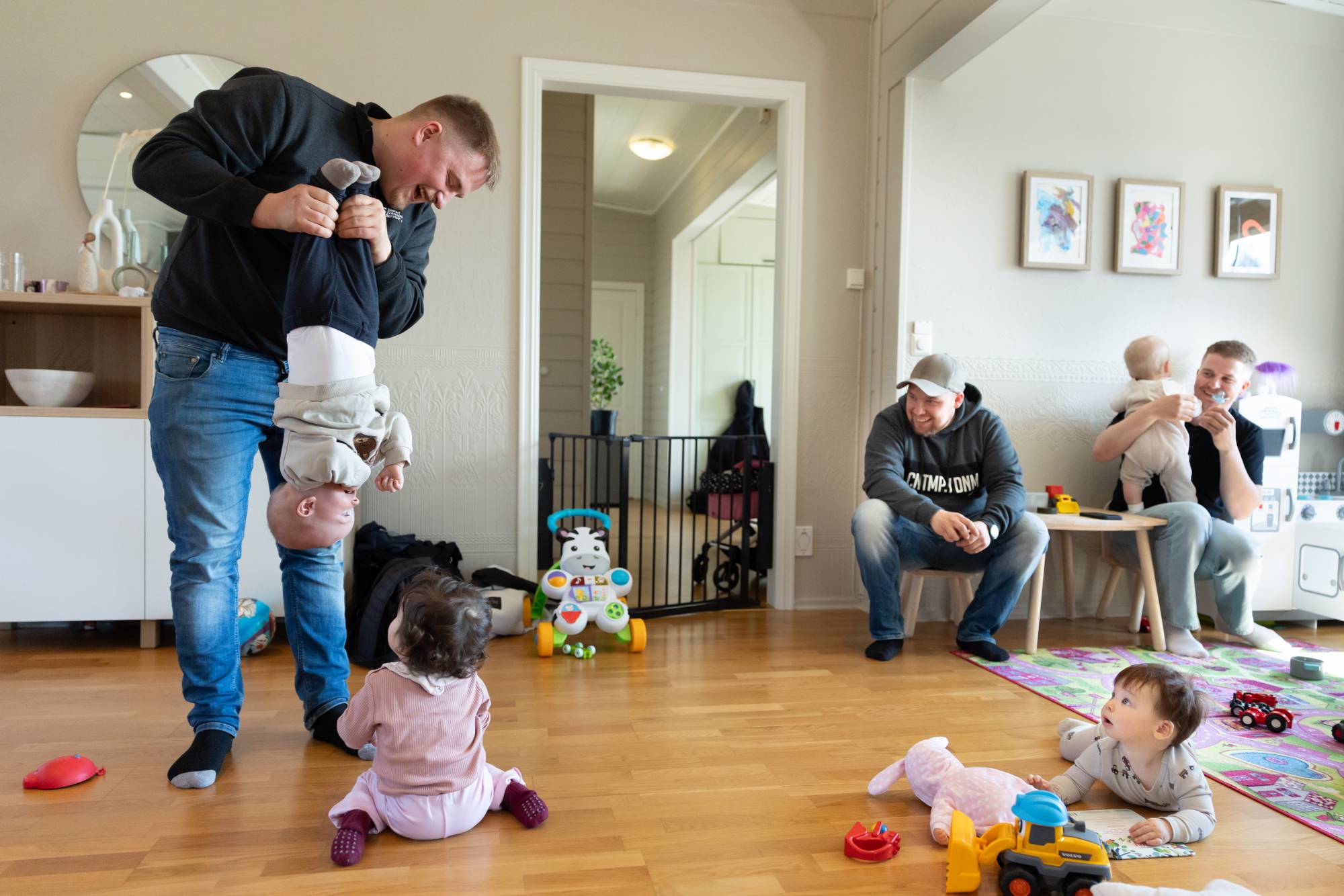 En mann holder en baby opp-ned, ansikt til ansikt mot en annen baby, mens to pappaer og en tredje baby ser på