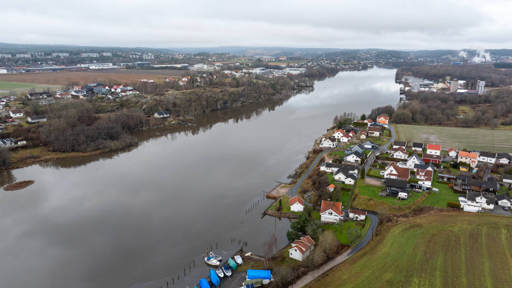 Bildet viser et panoramisk utsikt over en elv som renner gjennom et boligområde. På den ene siden av elven er det flere hus og båter, mens den andre siden har industri og grönområder i bakgrunnen. Landskapet er preget av høstfarger og er overskyet. Det er tydelige spor av bebyggelse og landbruk langs elven. (Bildebeskrivelsen er laget av en KI-tjeneste)