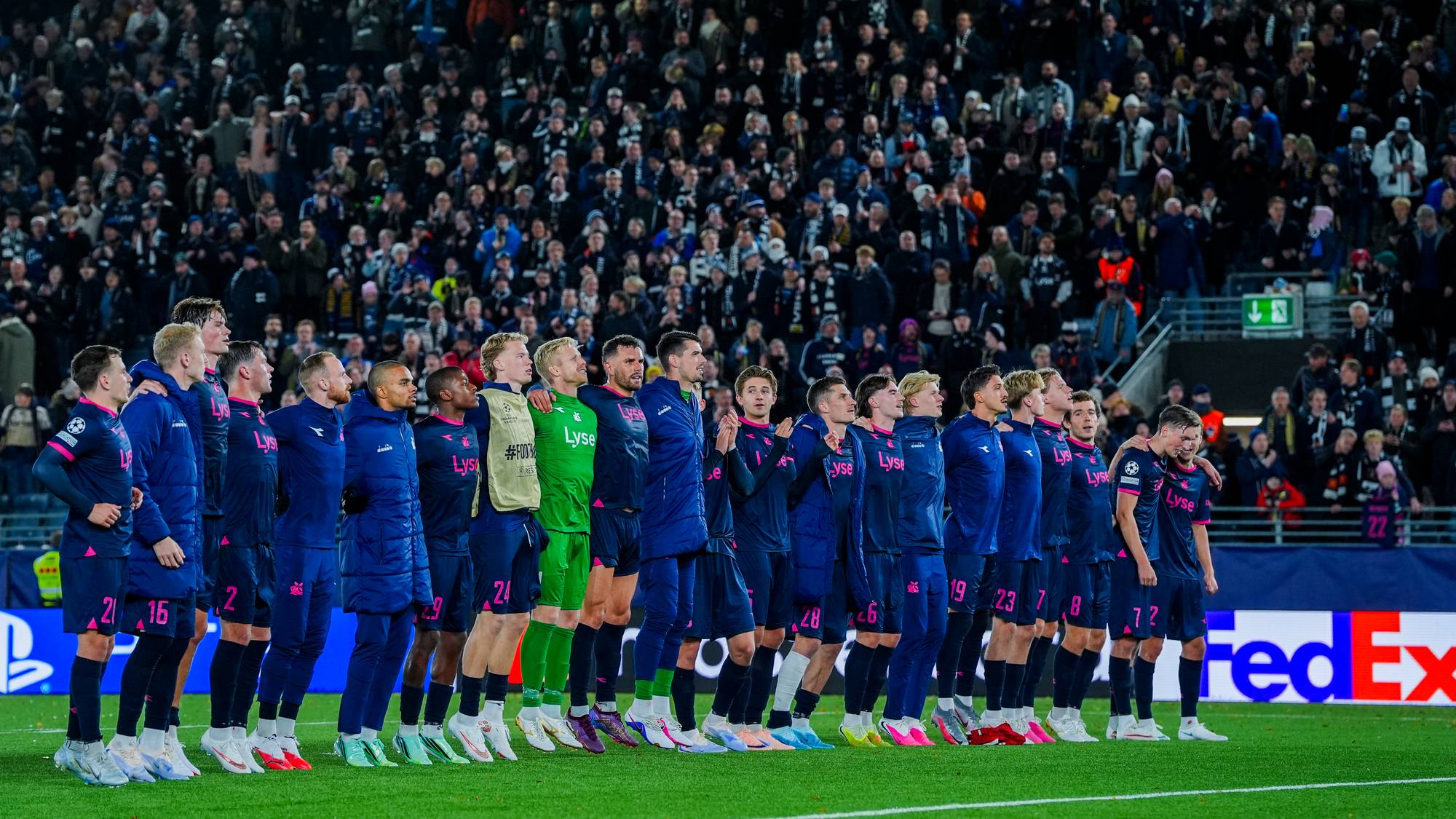 Fotballspillere står oppstilt på en rekke på gresset foran et fullsatt tribuneparti. (Bildebeskrivelsen er laget av en KI-tjeneste)