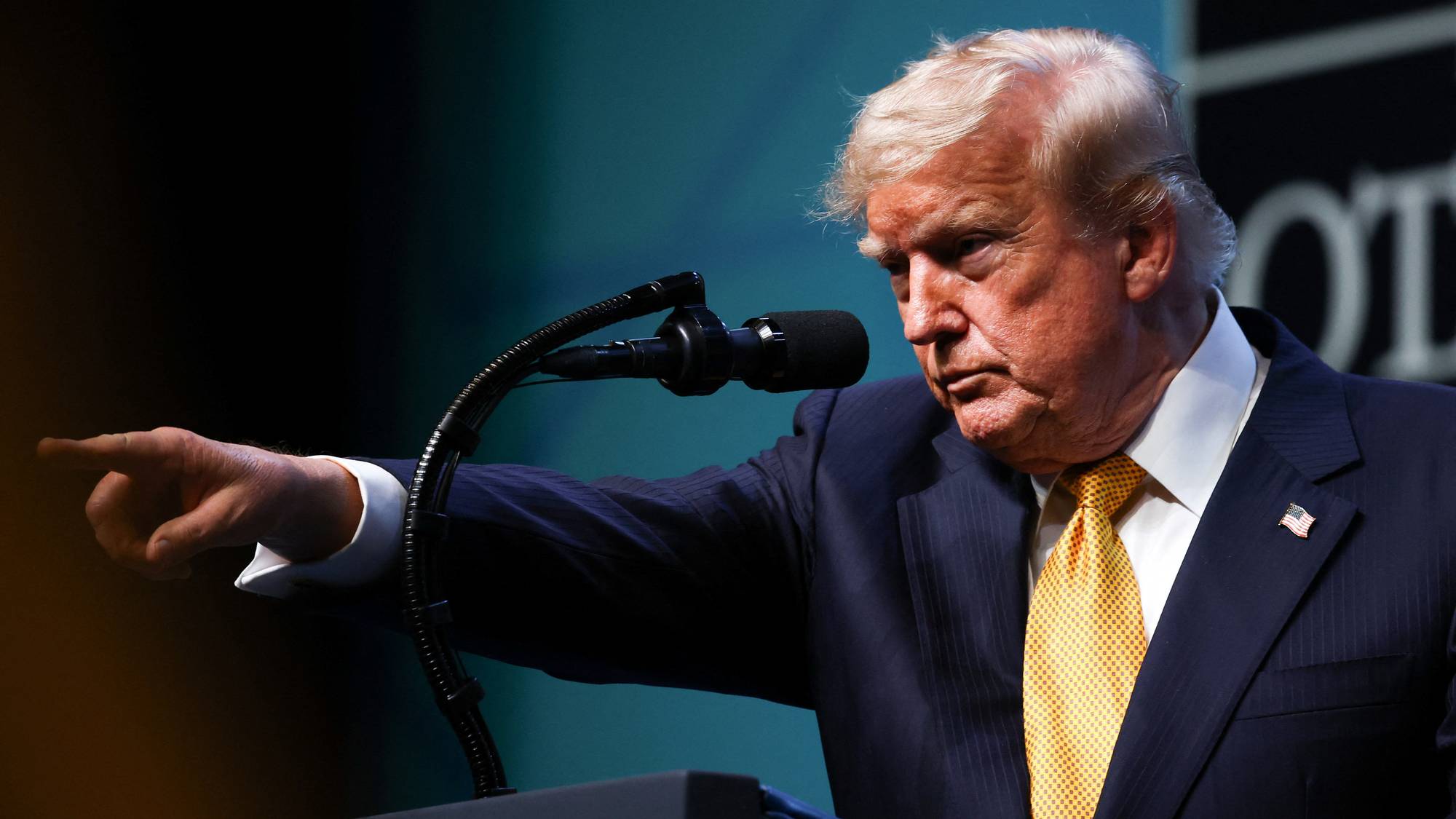 Donald Trump taler bak en mikrofon og peker ut til siden. Han er kledd i mørk dress og gult slips. 