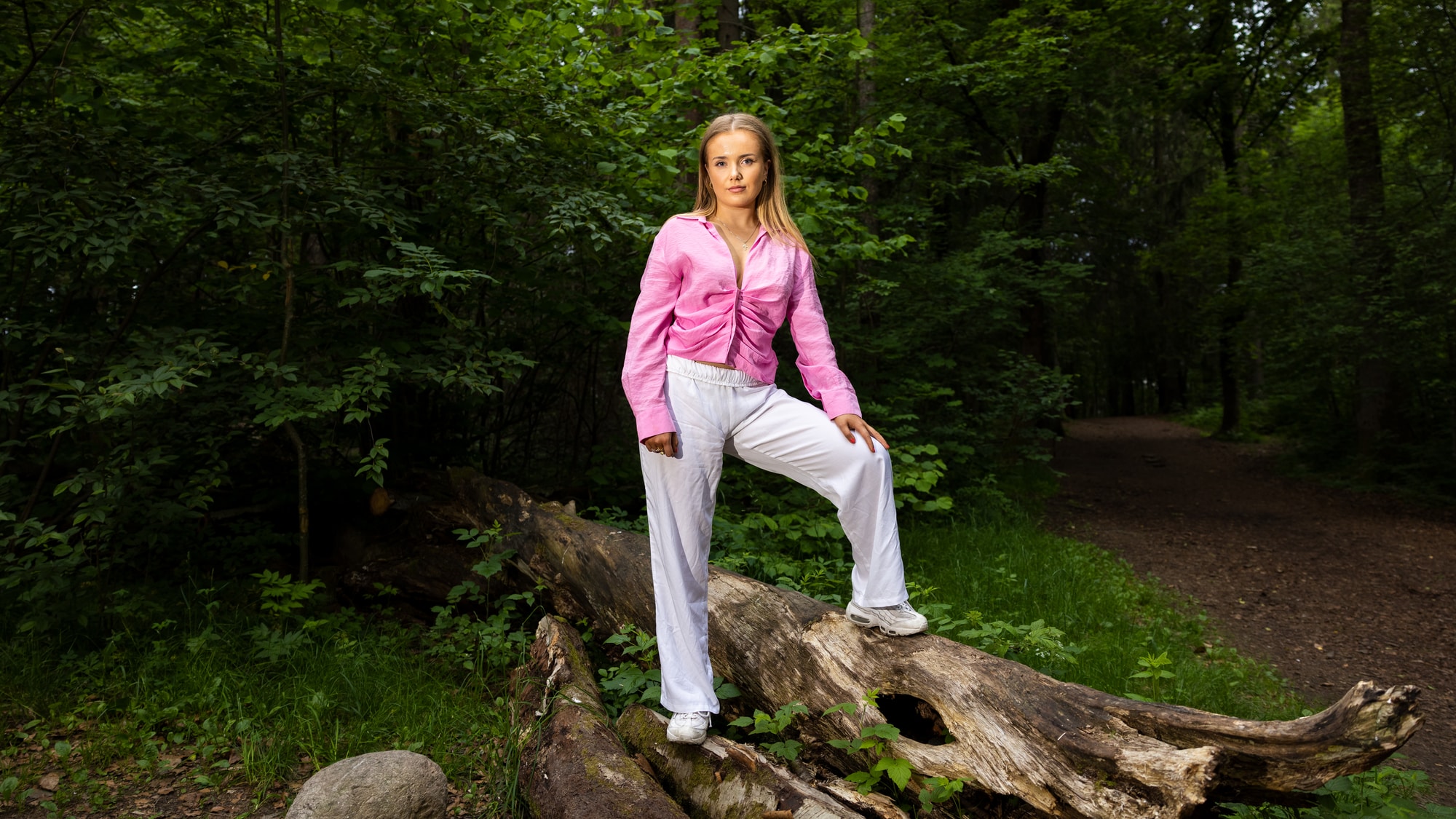 Bilde av Helena i rosa skjorte og hvit bukse stående med armene i kors på en trestubbe i Ekebergparken i Oslo. 