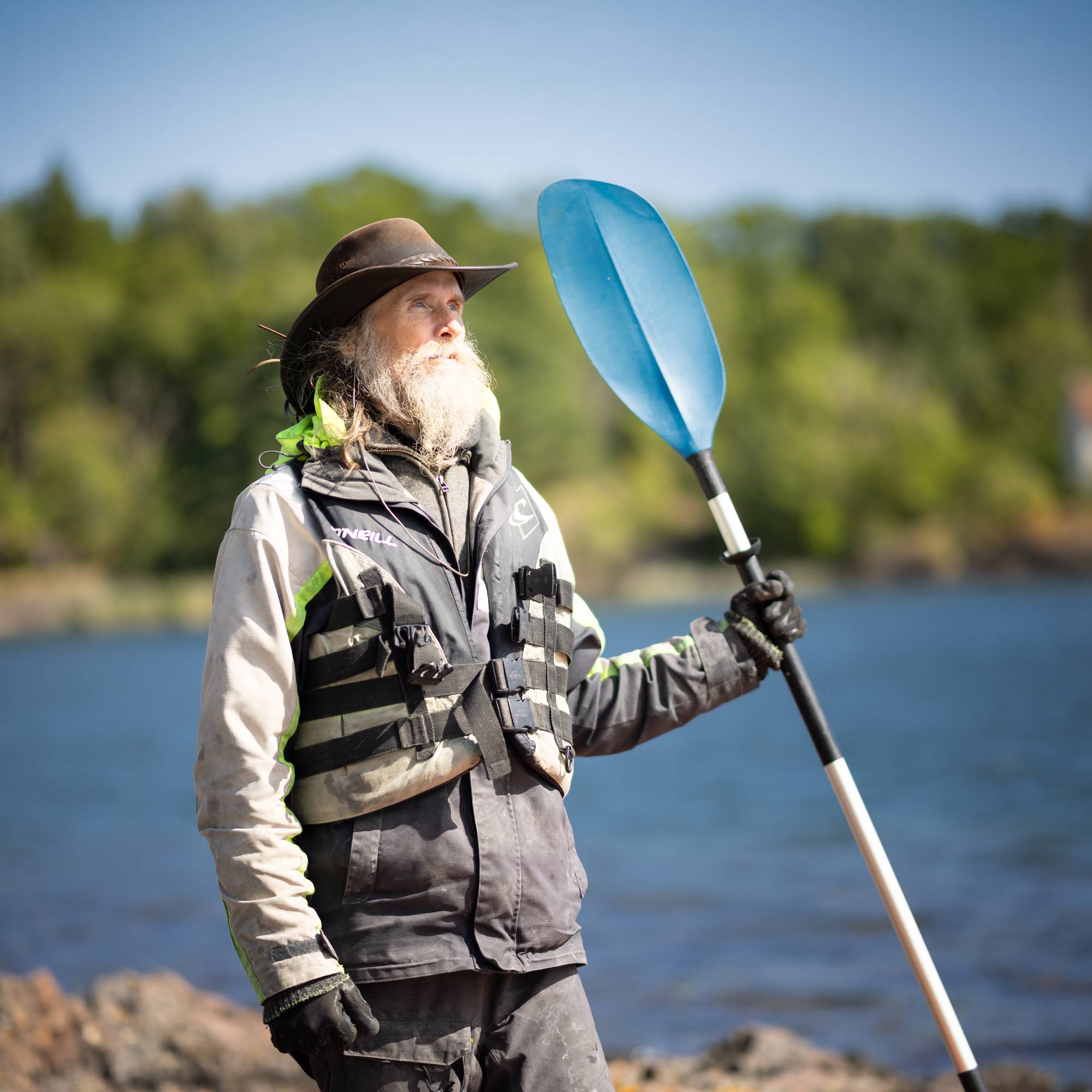 En mann med langt grått skjegg og hatt. Han har på seg redningsvest og holdere en åre i hånden. Han ser utover havet.