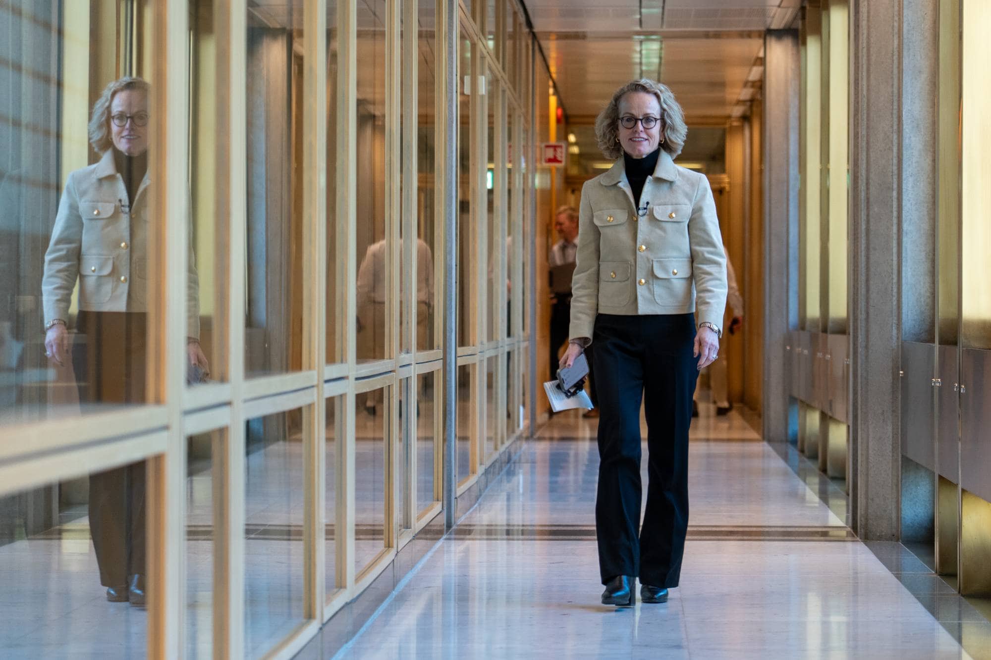 En kvinne med briller og krøllete hår går nedover en korridor. Hun har på seg en lys jakke og svarte bukser. Kvinnen holder en mobiltelefon og papirer i hånden. Bak henne sees andre personer og gjenskinnet av en annen person i glassveggen. (Bildebeskrivelsen er laget av en KI-tjeneste)