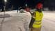 Ann-Kristin Hanssen / NRK En person i refleksvest og hjelm holder et lysende skilt. En bil står i veikanten i snø og mørke. En annen person er i ferd med å gå vekk fra bilen. Veien er snødekt og opplyst av lyktestolper. (Bildebeskrivelsen er laget av en KI-tjeneste)