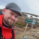 Erling Andre Solbakken Leine / Privat Portrettbilde av en mann som står på et fjell med natur i bakgrunnen og et turstiskilt ved siden av seg.