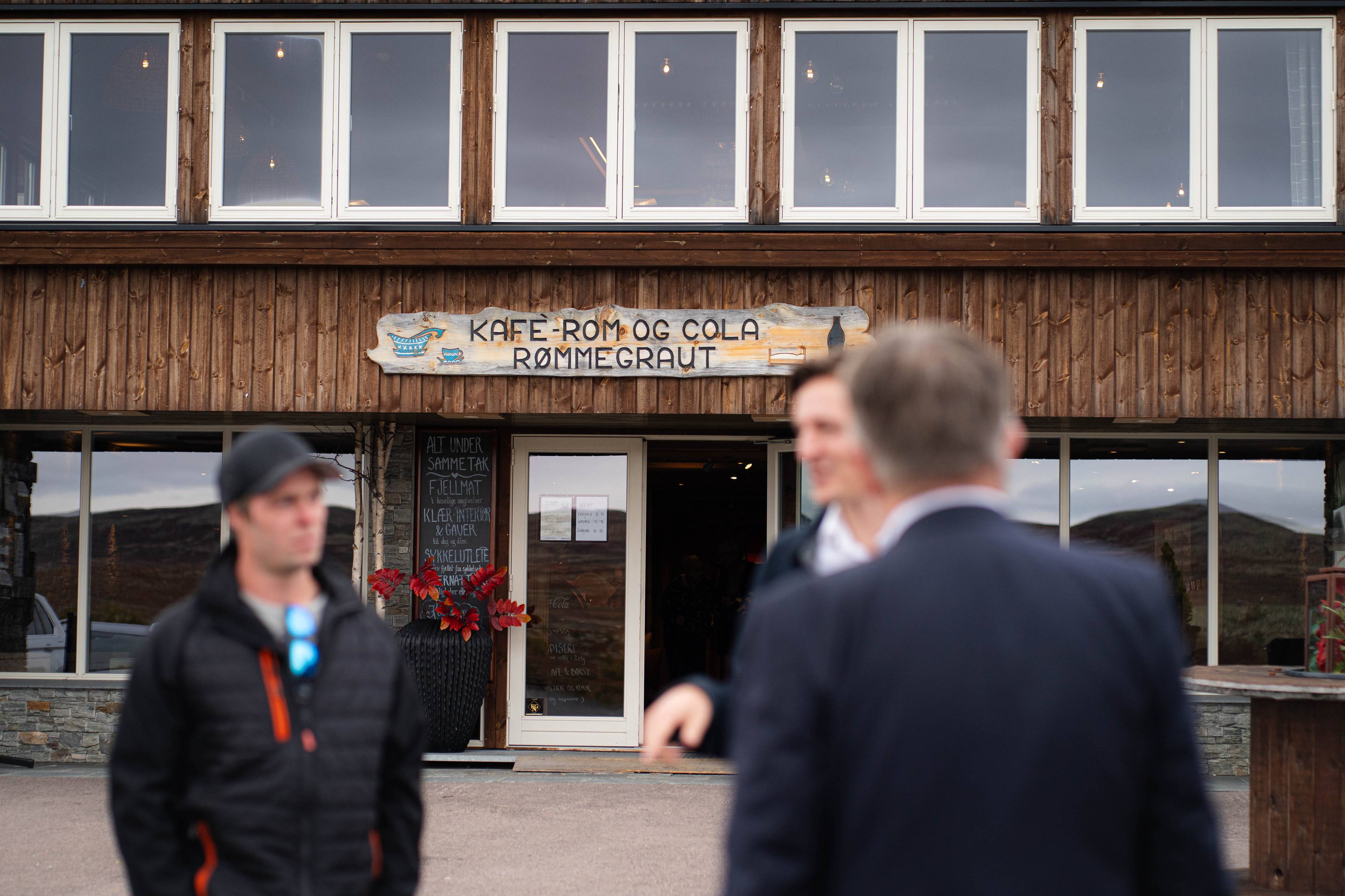 Personer foran en bygning med skiltet Kafe-rom og cola rømmegraut. (Bildebeskrivelsen er laget av en KI-tjeneste)