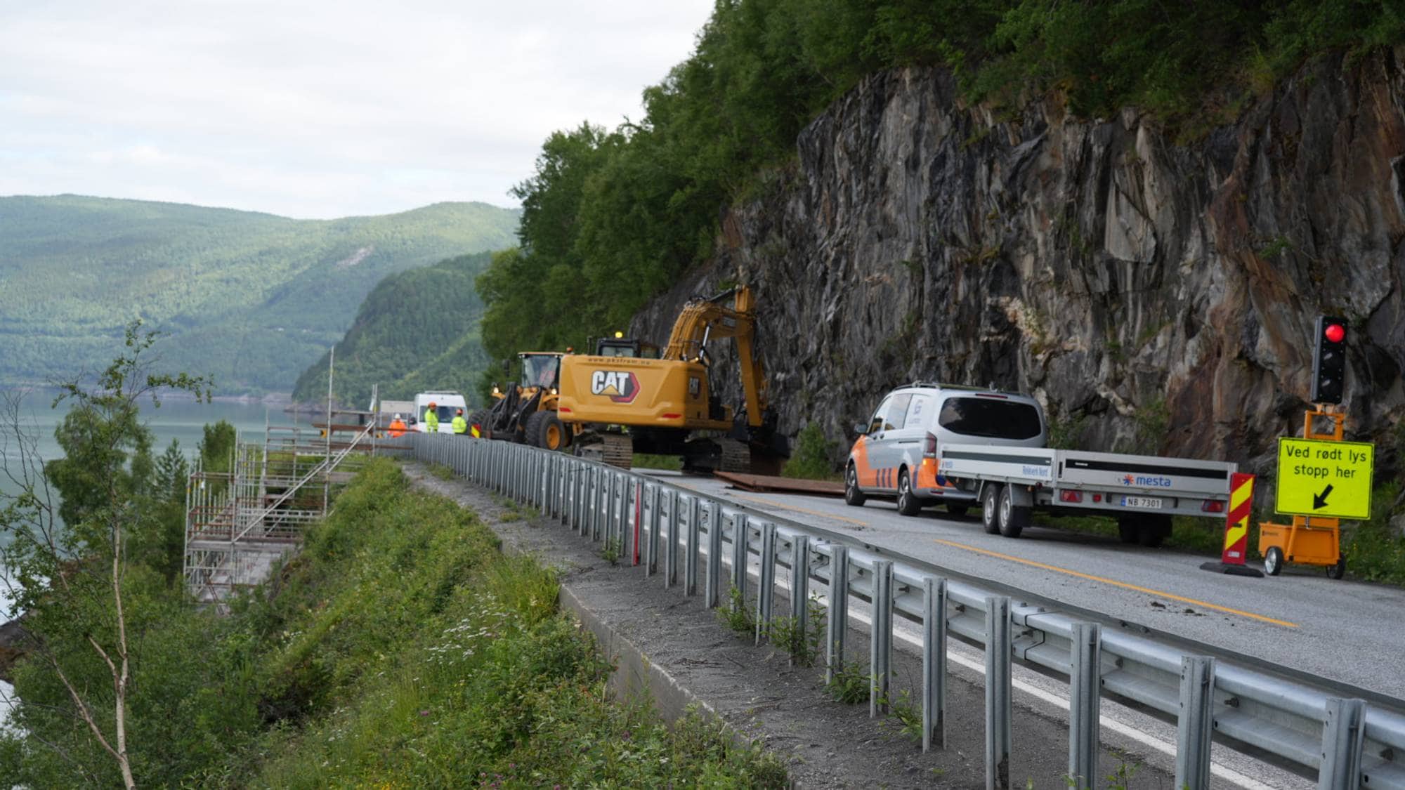 Veiarbeid med gravemaskin og kjøretøy langs fjellvei ved en innsjø. Trafikklys viser rødt. (Bildebeskrivelsen er laget av en KI-tjeneste)