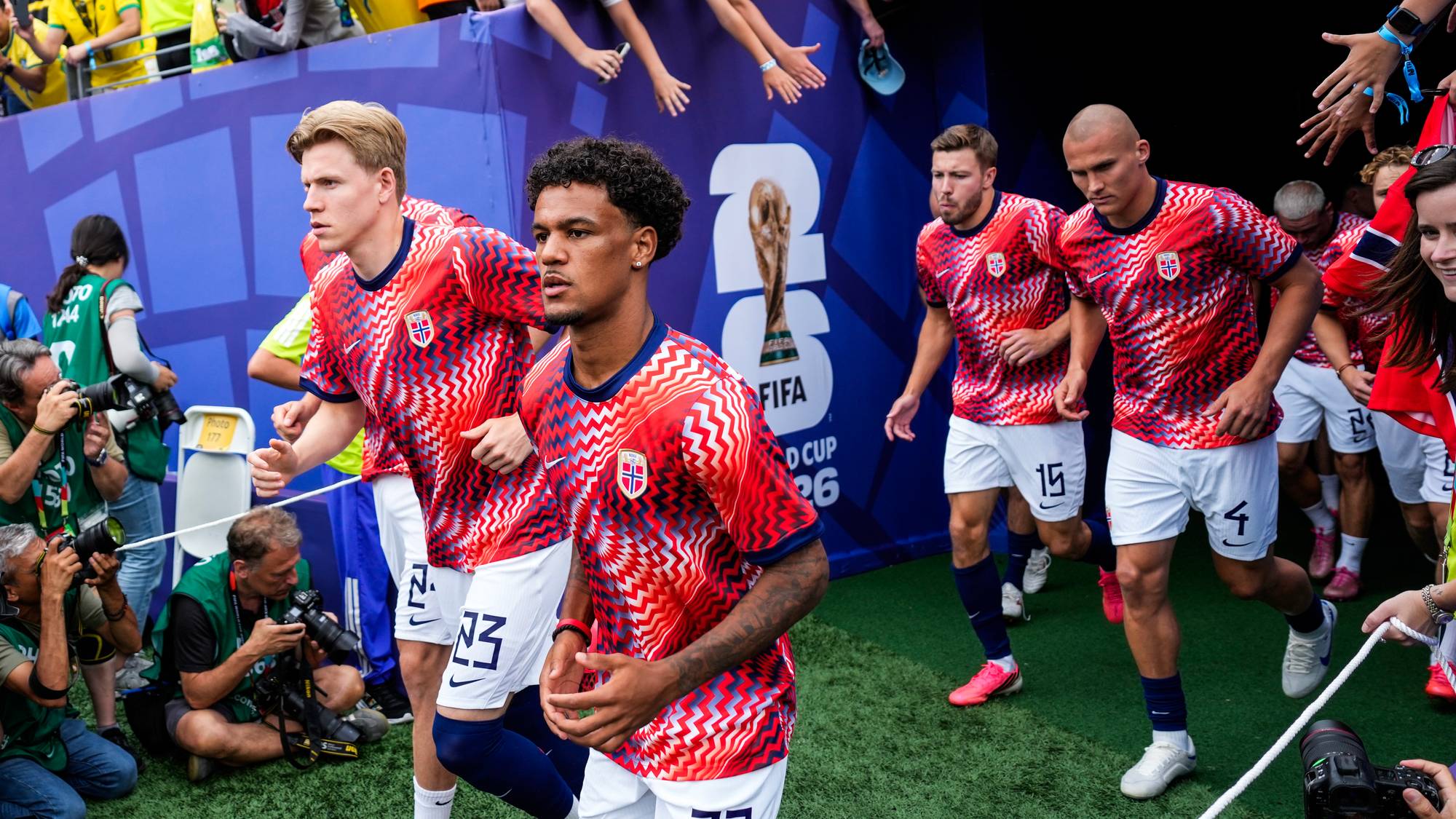 Norske fotballspillere i røde drakter løper ut på banen foran et publikum på en stadion. (Bildebeskrivelsen er laget av en KI-tjeneste)