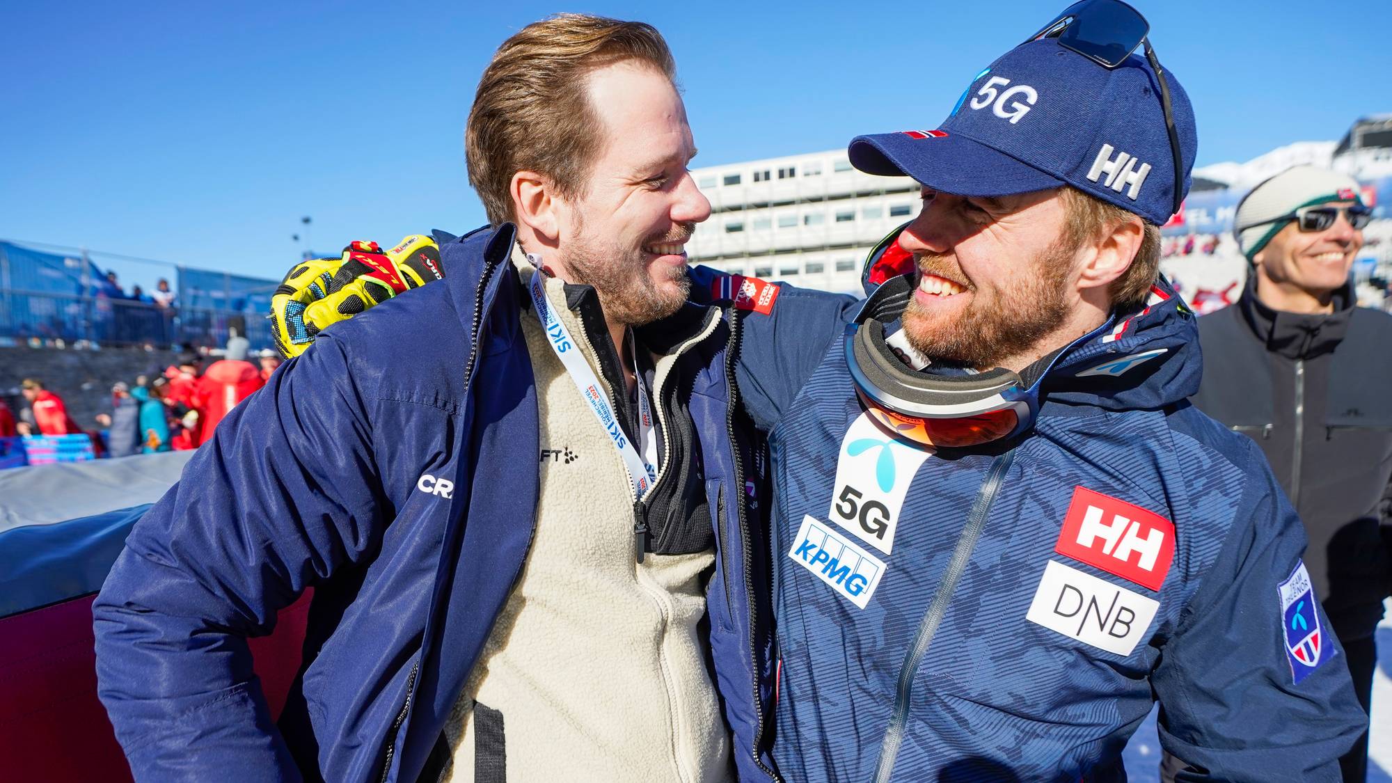 To menn i skiklær står sammen og smiler utendørs i solfylt vintervær. (Bildebeskrivelsen er laget av en KI-tjeneste)