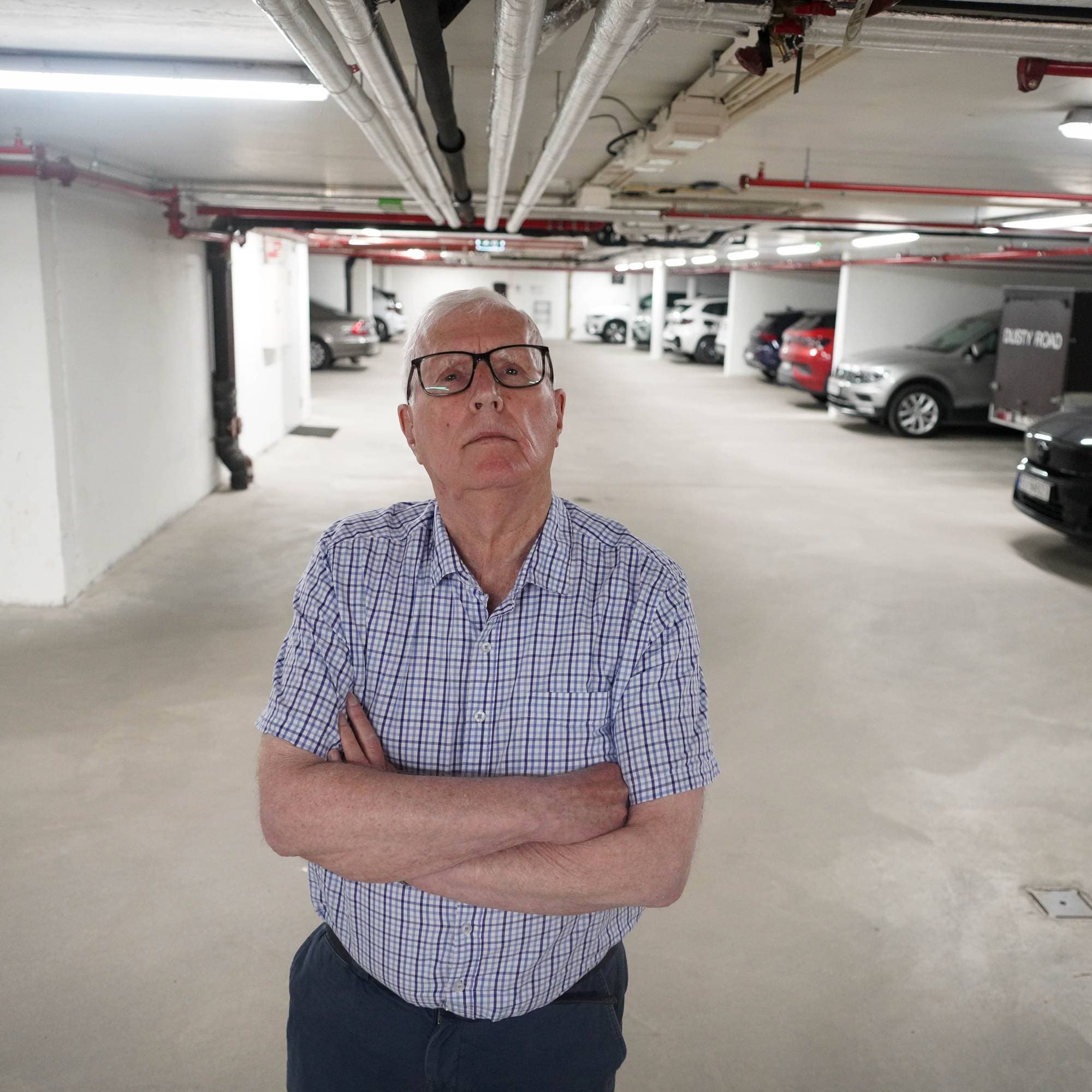 En eldre mann med briller og rutete skjorte står med armene i kors. Han står i en parkeringskjeller med flere biler og lysstoffrør i taket. Bak mannen står en rekke biler parkert på rekke og rad. (Bildebeskrivelsen er laget av en KI-tjeneste)