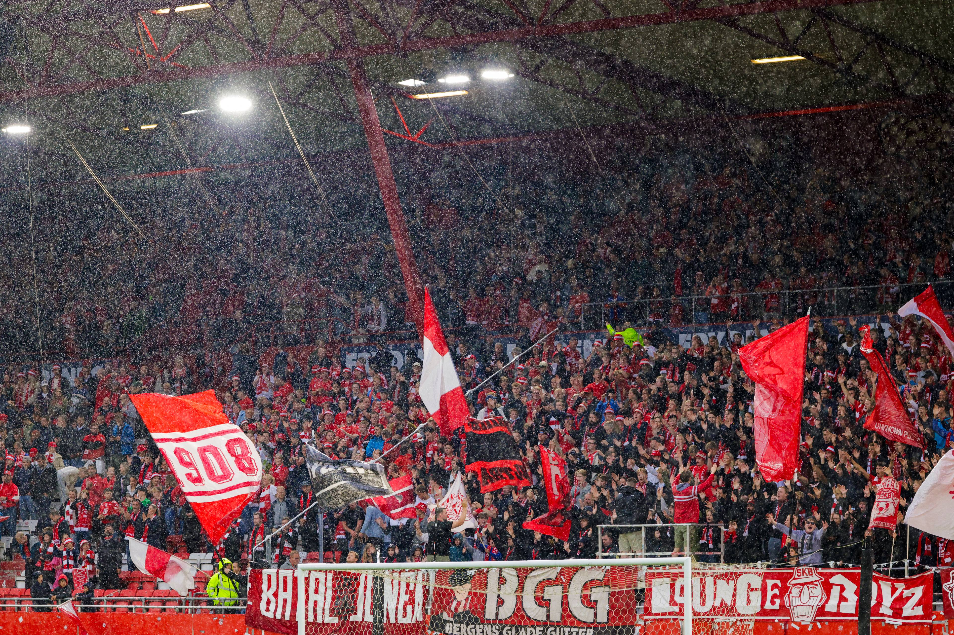 Supportere på en tribunetribune under en fotballkamp i regnvær. Mange vifter med røde flagg. (Bildebeskrivelsen er laget av en KI-tjeneste)