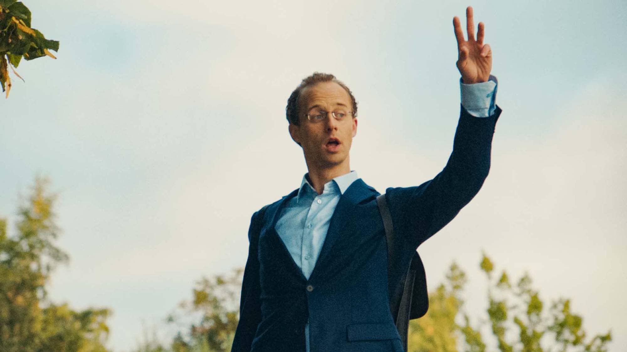 Fridtjof Josefsen har på seg dress, blå skjorte og briller. Han står ute blant trær og vinker. Ansiktsuttrykket hans er nøytralt.