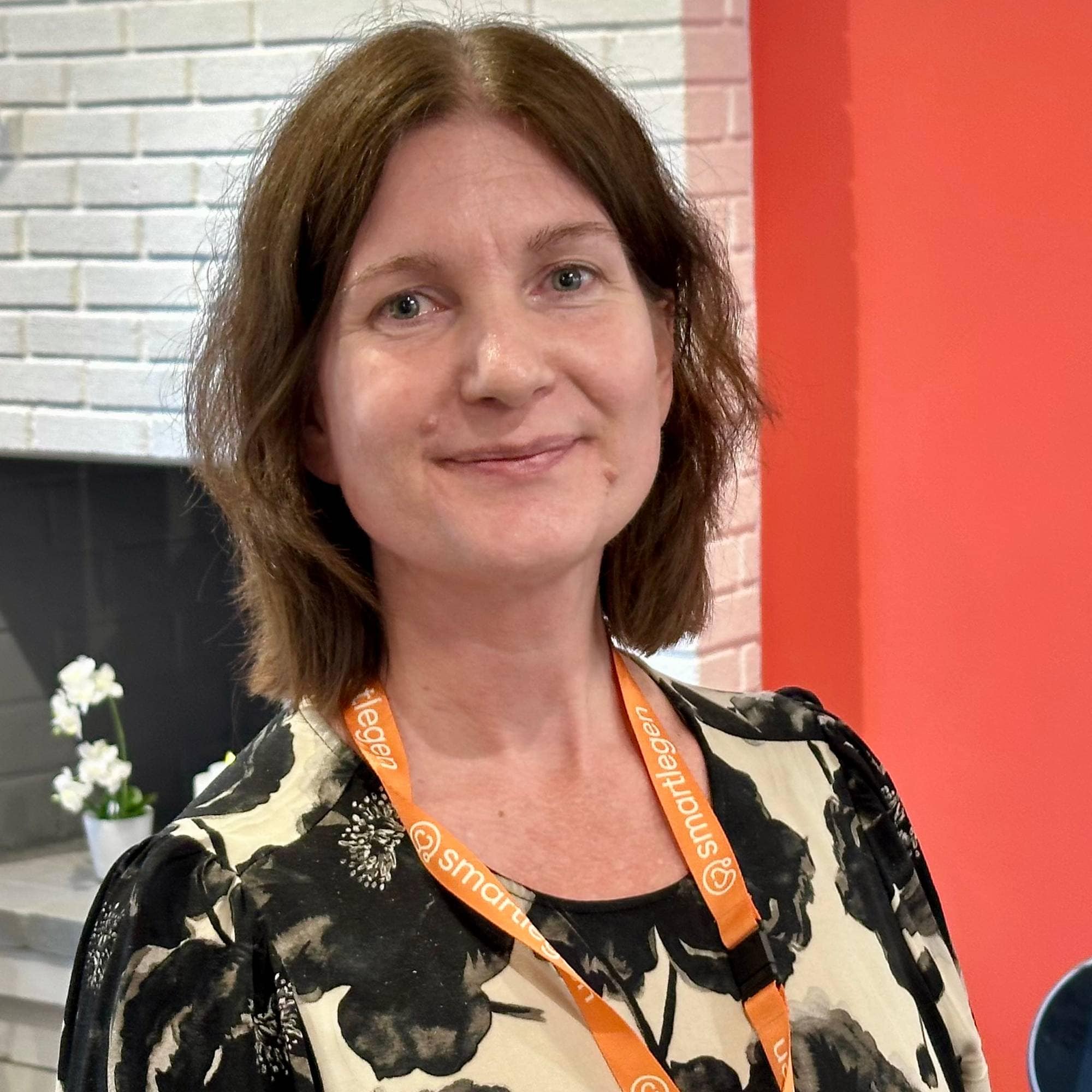 Kvinne med mønstret bluse og oransje bånd rundt halsen står foran en hvit murvegg. (Bildebeskrivelsen er laget av en KI-tjeneste)