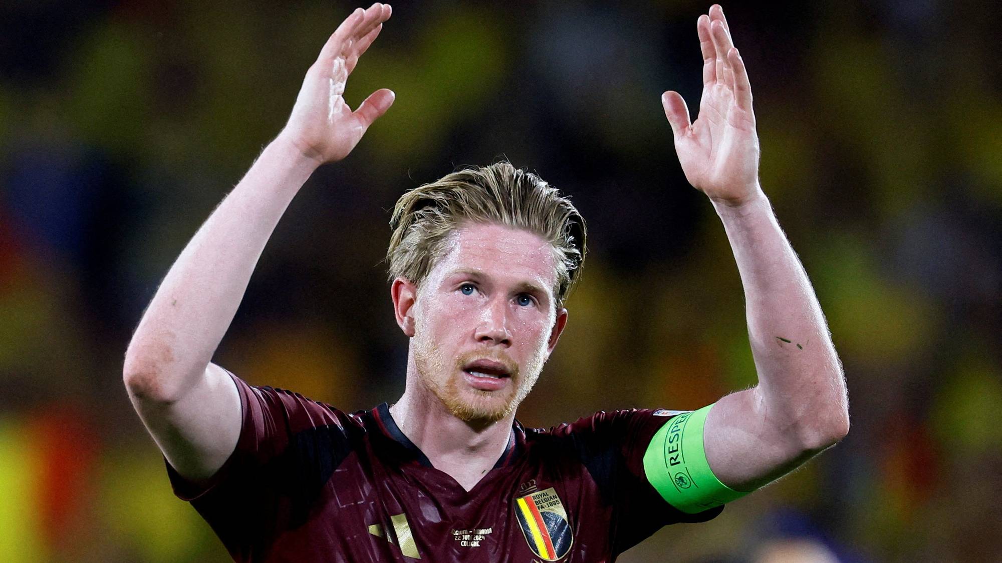 En fotballspiller i mørkerød drakt med et hvitt og grønt bånd rundt venstre arm hever begge hender over hodet. Spilleren har blondt hår og en lys hudtone. I bakgrunnen er det uskarpe, gule og mørke farger. (Bildebeskrivelsen er laget av en KI-tjeneste)