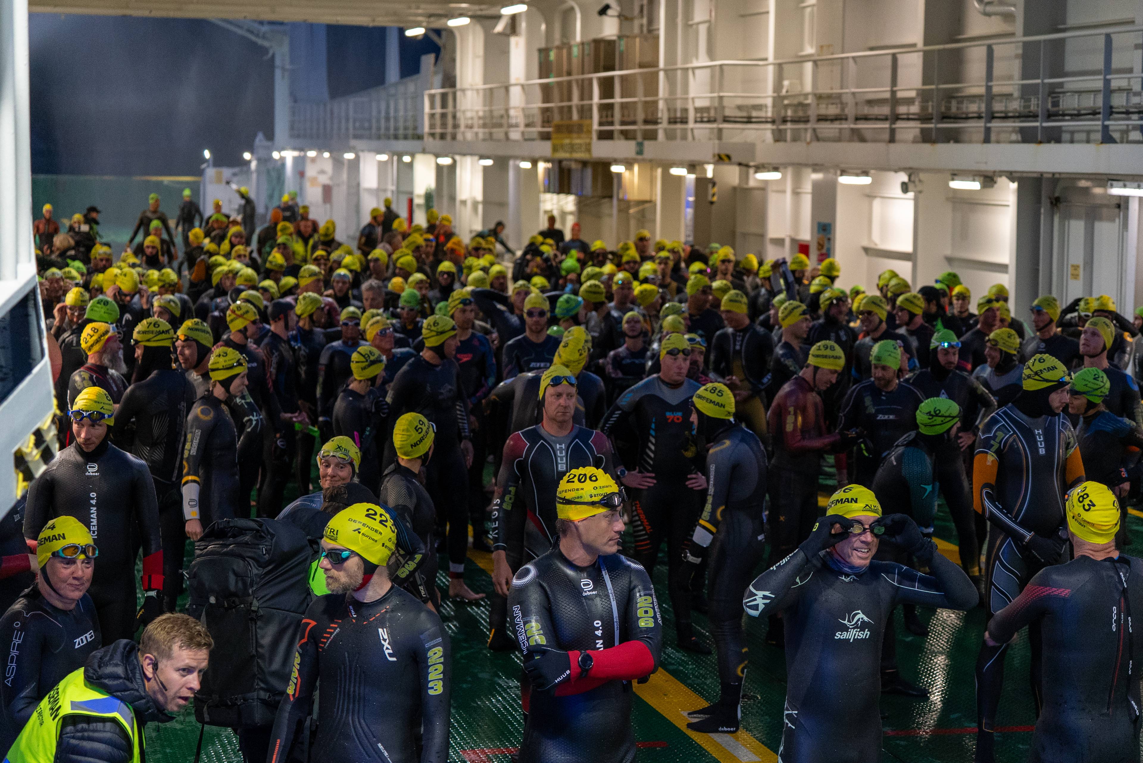 Deltakere i våtdrakt og gul badehette samlet på et fergedekk før Norseman-triatlon. (Bildebeskrivelsen er laget av en KI-tjeneste)